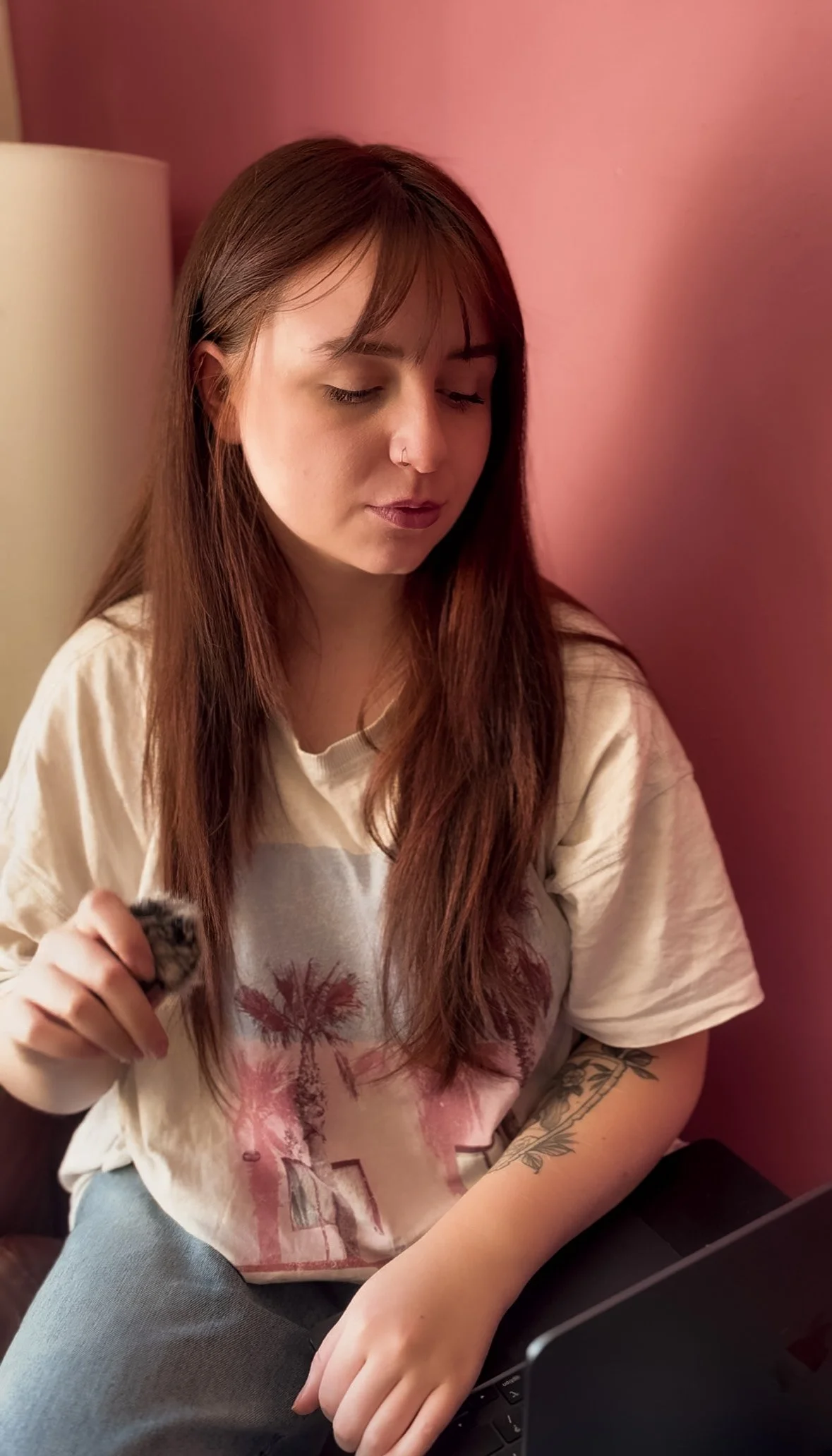 Young woman with long brown hair and a nose ring sitting against a pink wall, wearing a beige T-shirt with palm trees, working on a laptop and holding a small wireless microphone