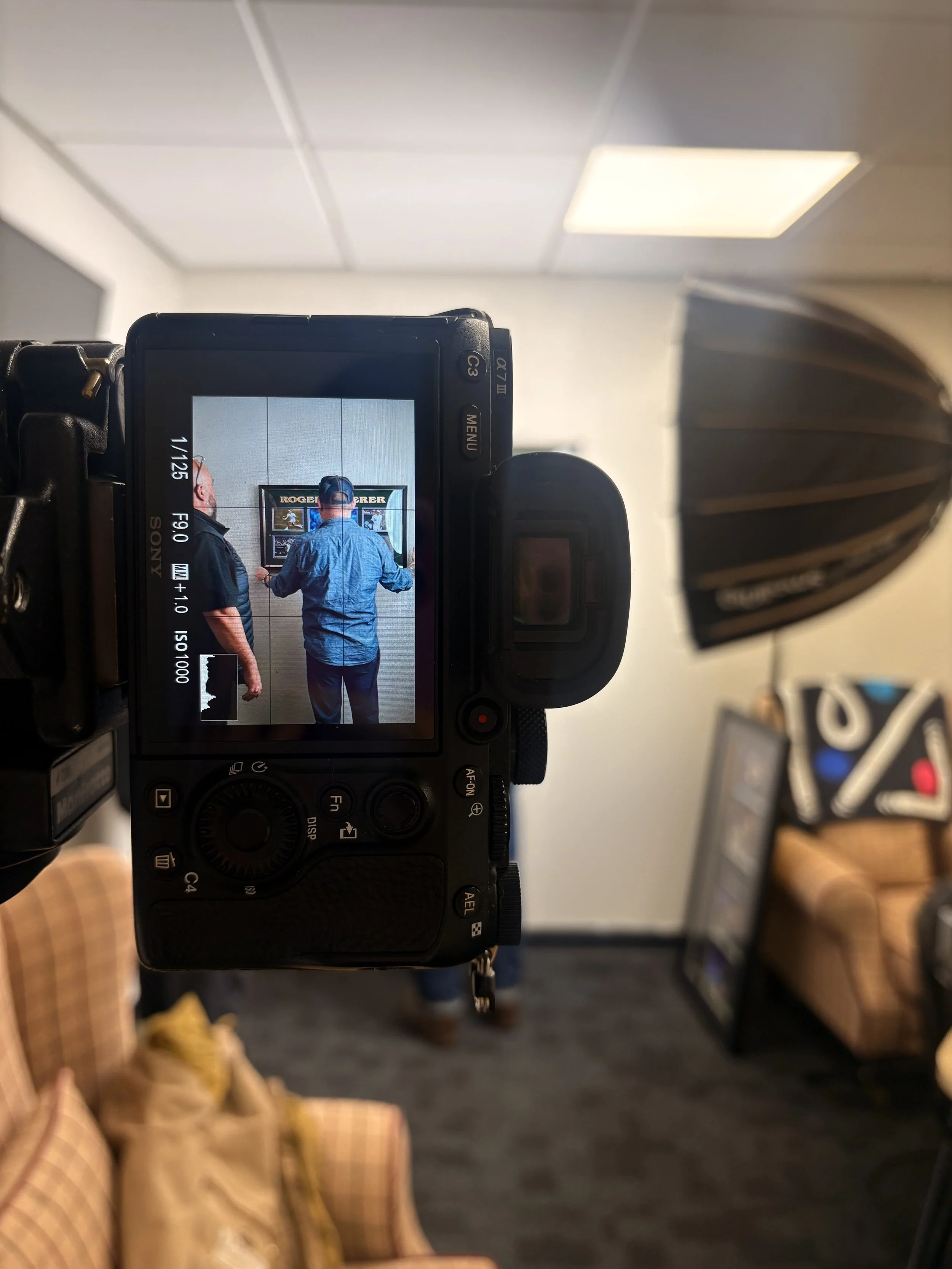 Camera capturing a photo of two men standing in front of a wall display with framed pictures, one with a hat and the other with short hair, inside a room with white ceiling tiles and a dark carpeted floor.