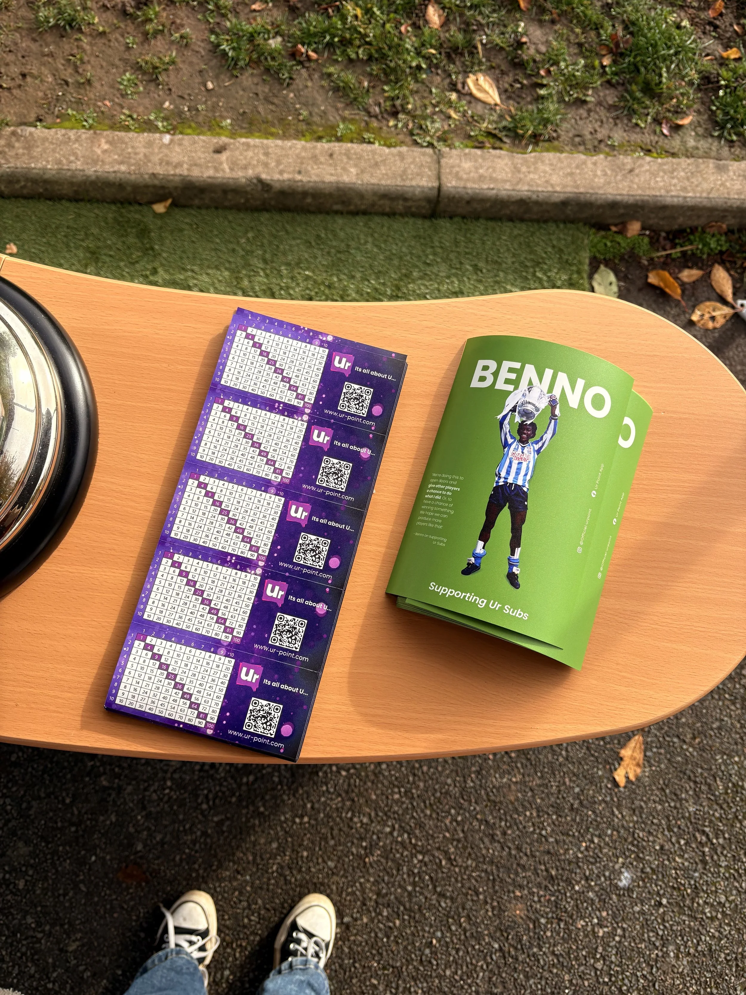 View of four lottery tickets on a wooden surface at outdoor setting, with a green card supporting a soccer player, and part of the photographer's shoes and jeans visible on asphalt ground.