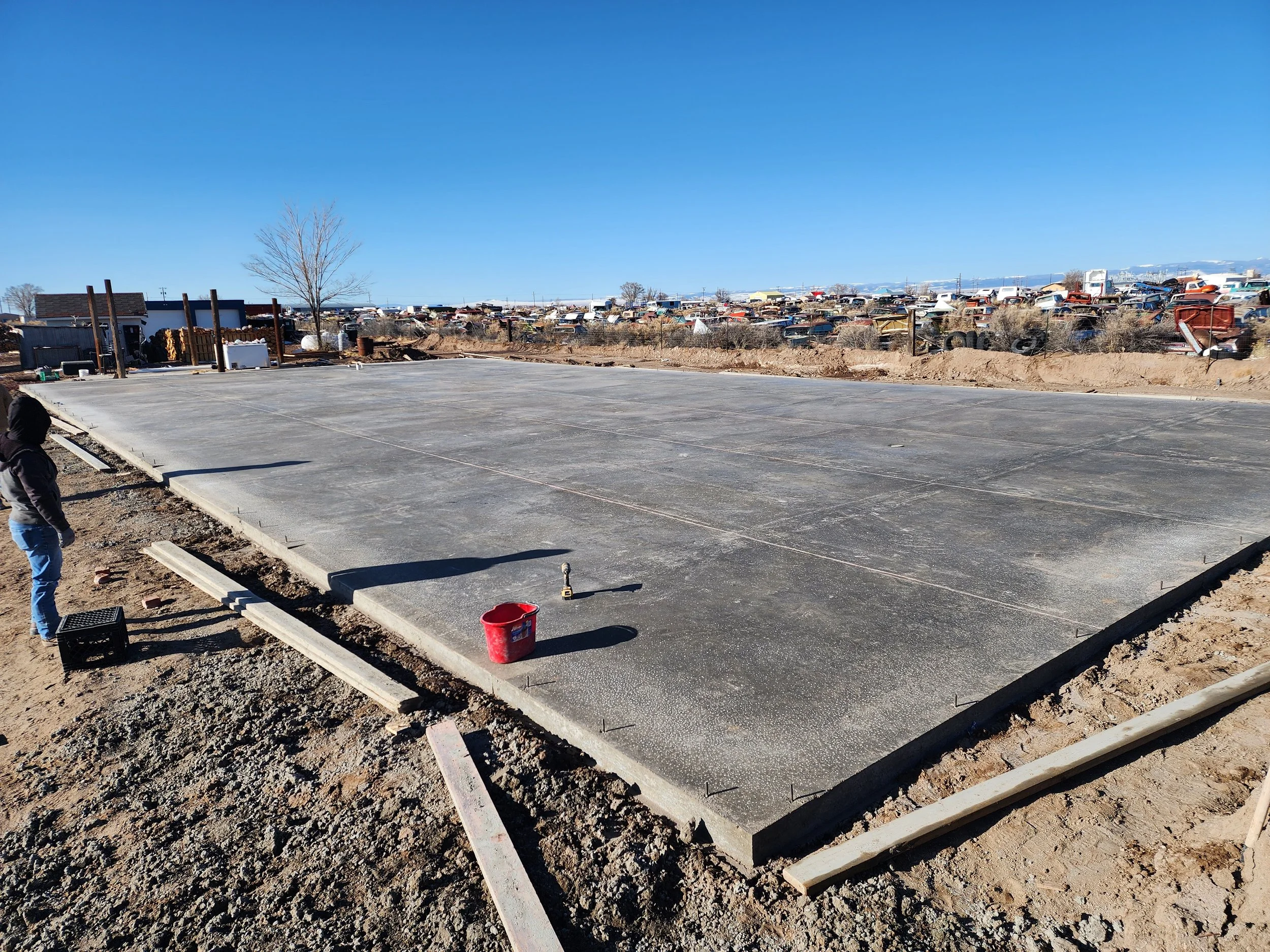 Freshly poured concrete slab on a construction site with a person standing nearby, a red bucket, and a clear blue sky.
