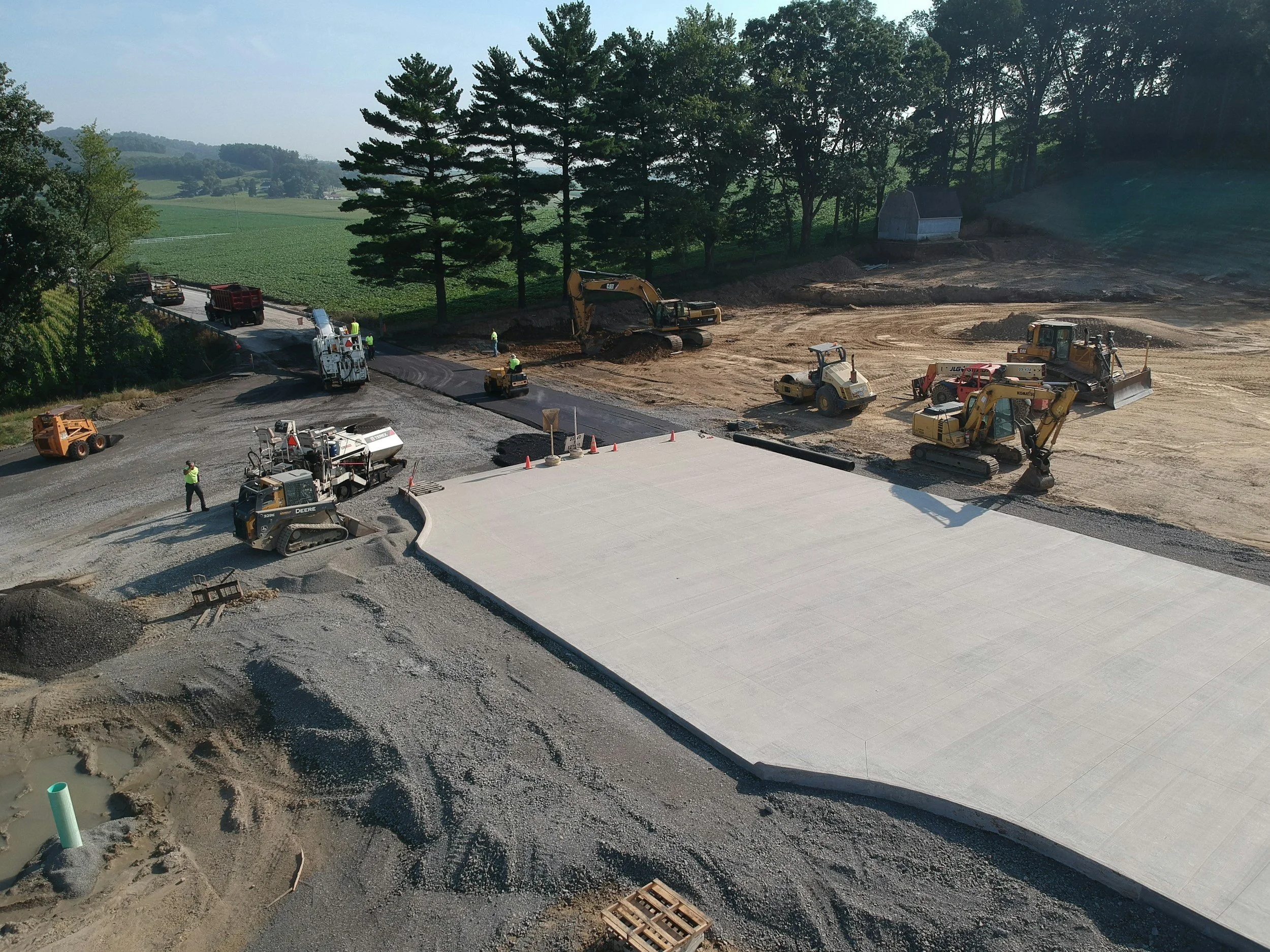 Construction site with multiple construction vehicles, workers, and freshly poured concrete on a road or parking lot under construction, with green fields and trees in the background.