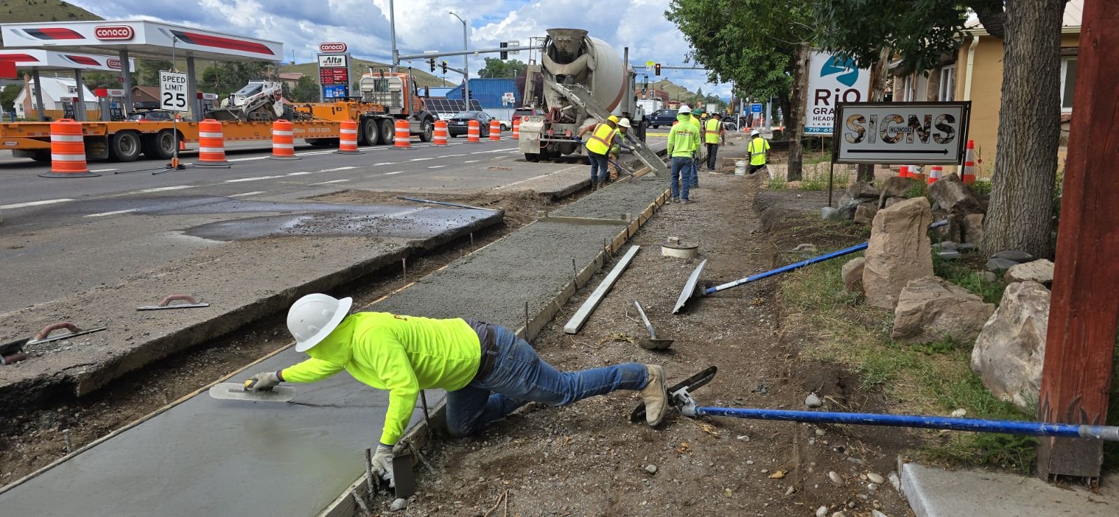 Construction workers in safety vests and helmets pouring and leveling concrete for a sidewalk along a busy street, with orange traffic cones and a cement mixer truck in the background.