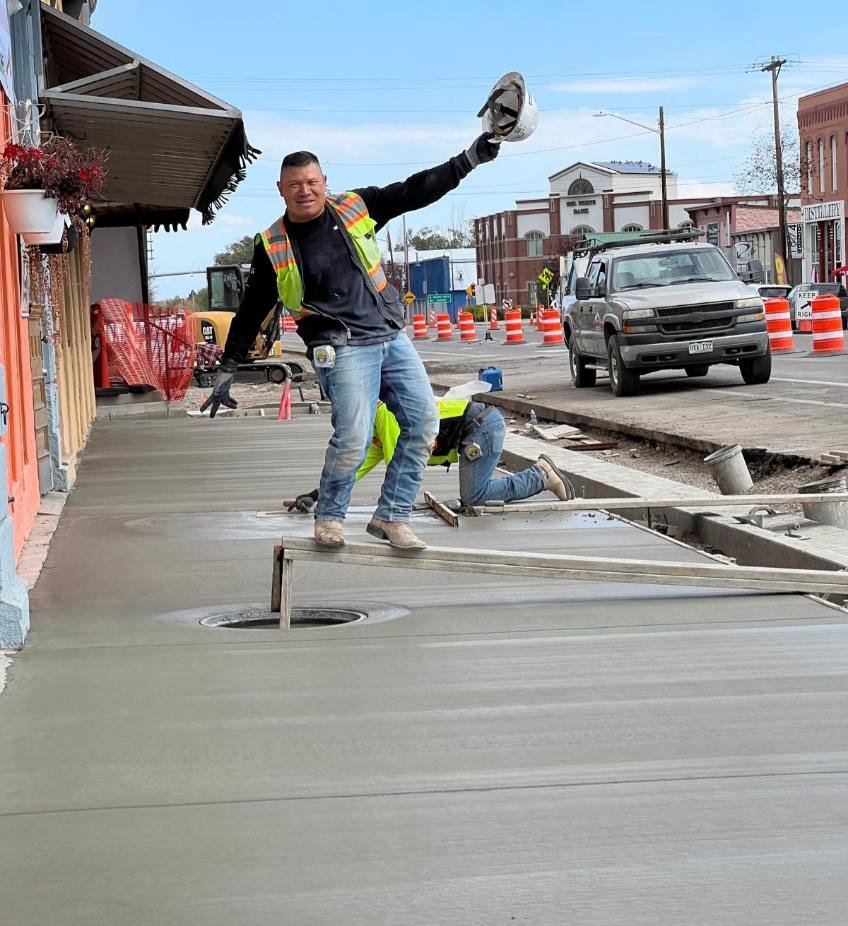 Construction workers pouring and finishing concrete sidewalk, one worker smiling and holding a construction helmet, another worker kneeling down working on the concrete, in a street with orange traffic cones and a parked vehicle.