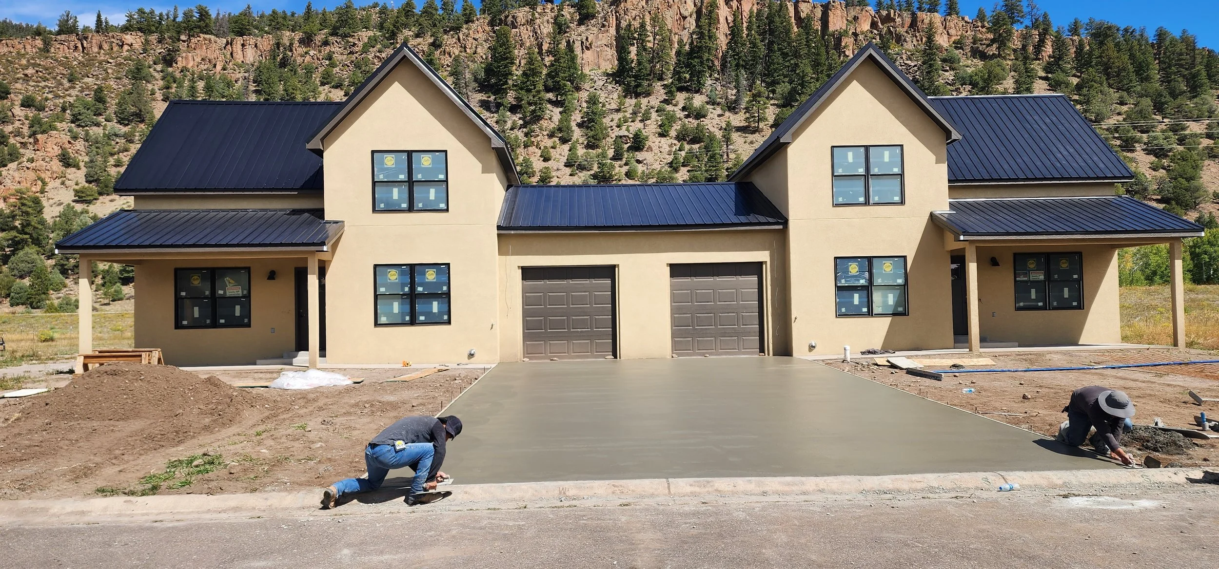 Construction workers are smoothing wet concrete on a driveway in front of a house under construction, with a mountainous landscape in the background.