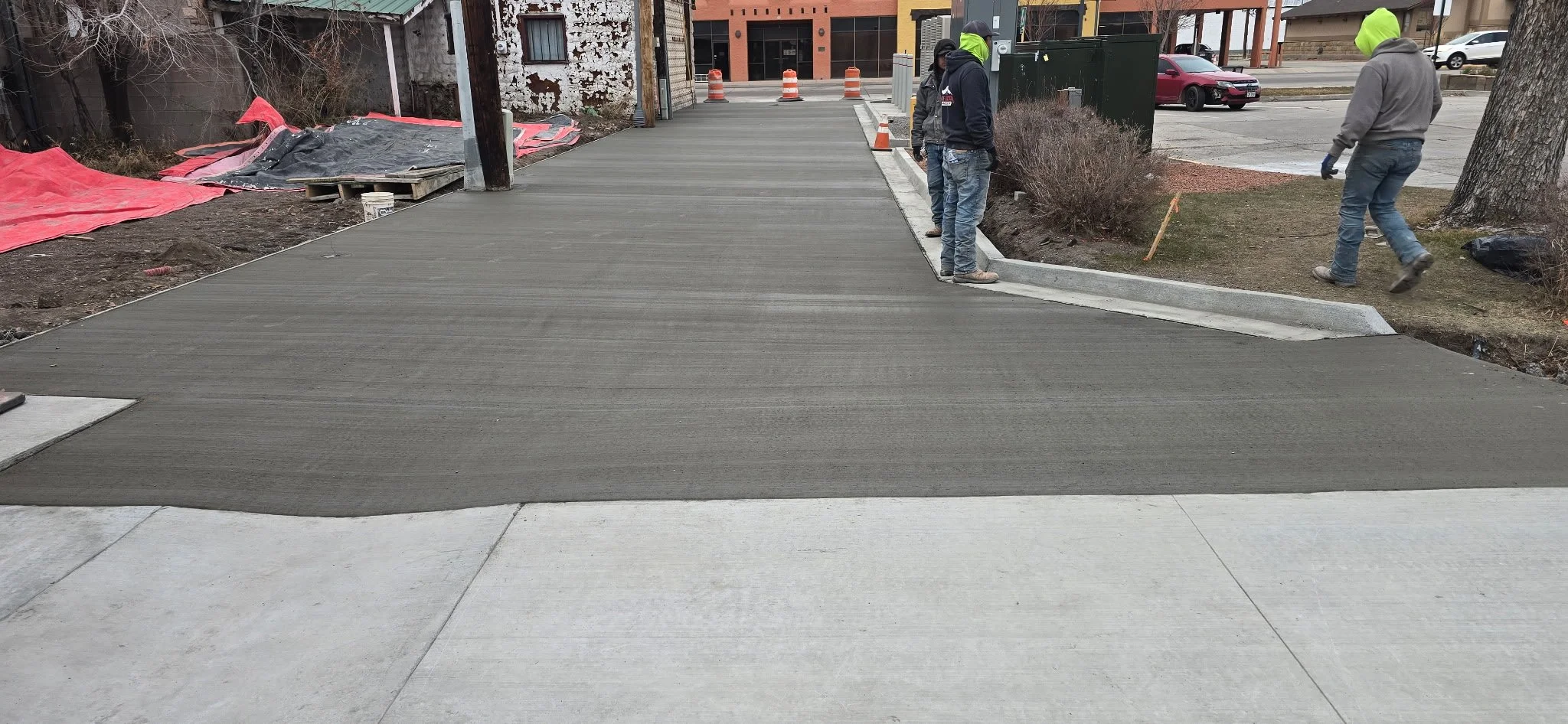 Workers are laying down fresh concrete on a sidewalk, with some standing near the edge, wearing work gloves and bright green hoodie caps. The freshly poured concrete is darker in color, contrasting with the light-colored existing sidewalk. Constructi