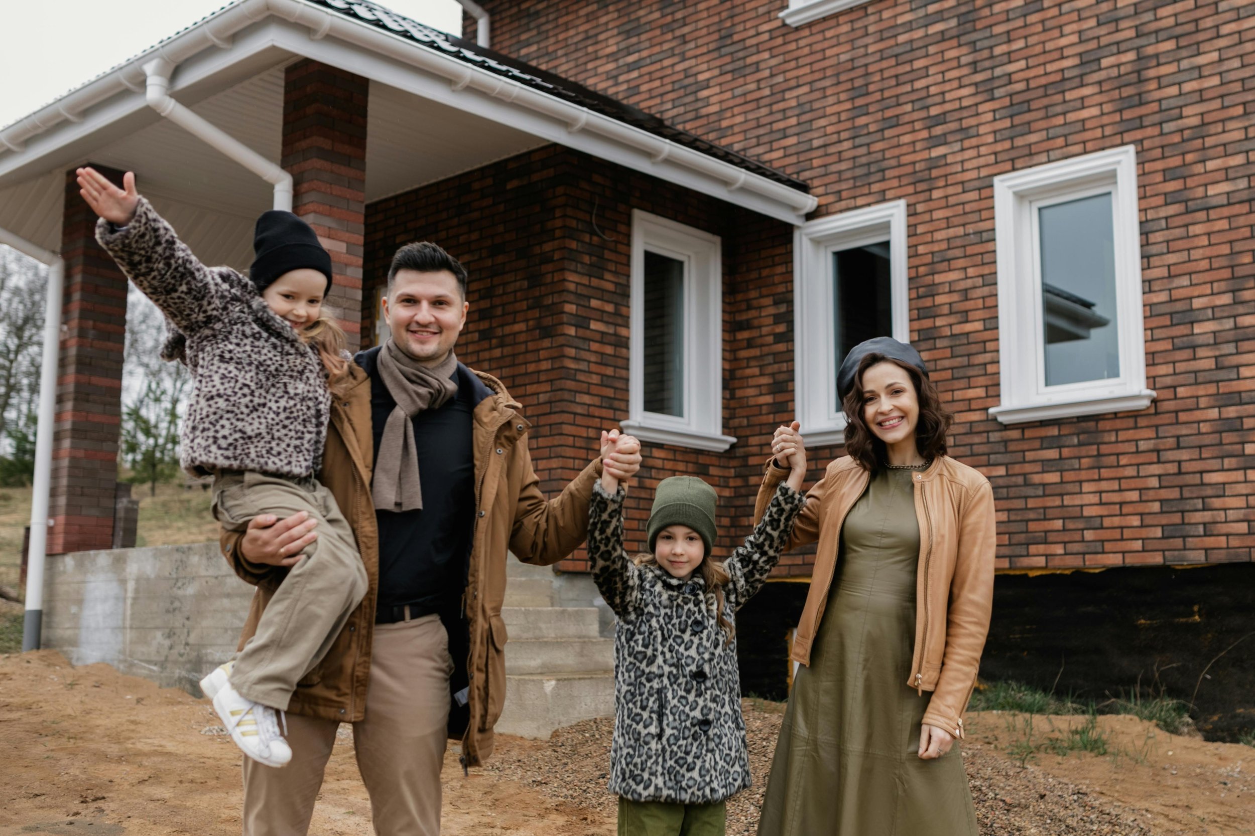 A family of five stands outside a brick house, holding hands and smiling.