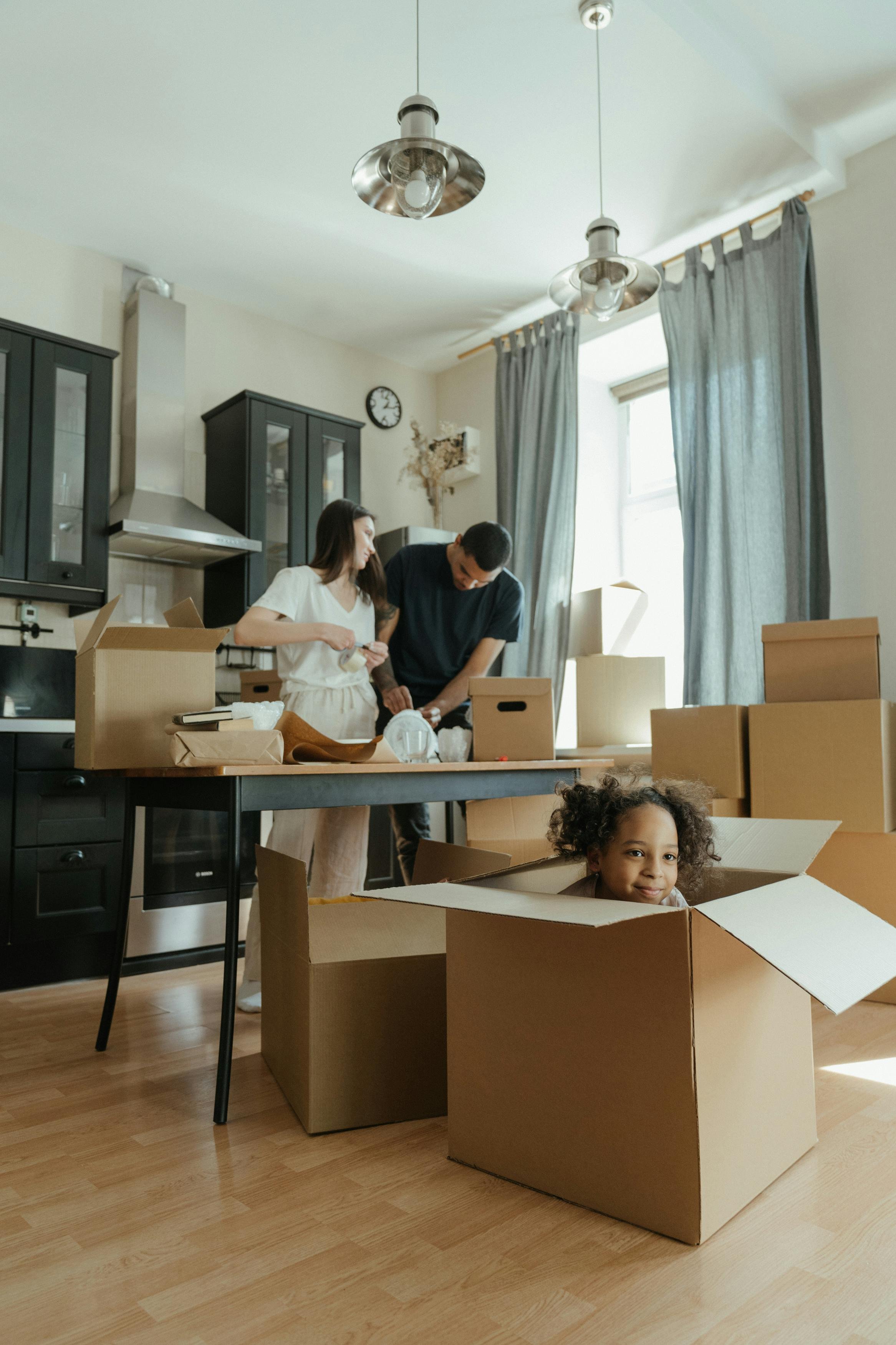 Two adults and a child are unpacking moving boxes in a bright kitchen with black cabinets and gray curtains. The child is sitting inside a large cardboard box, smiling at the camera. The adults are working together, with one woman holding a small box and a man leaning over a box on the table. The scene has natural sunlight coming through a window.