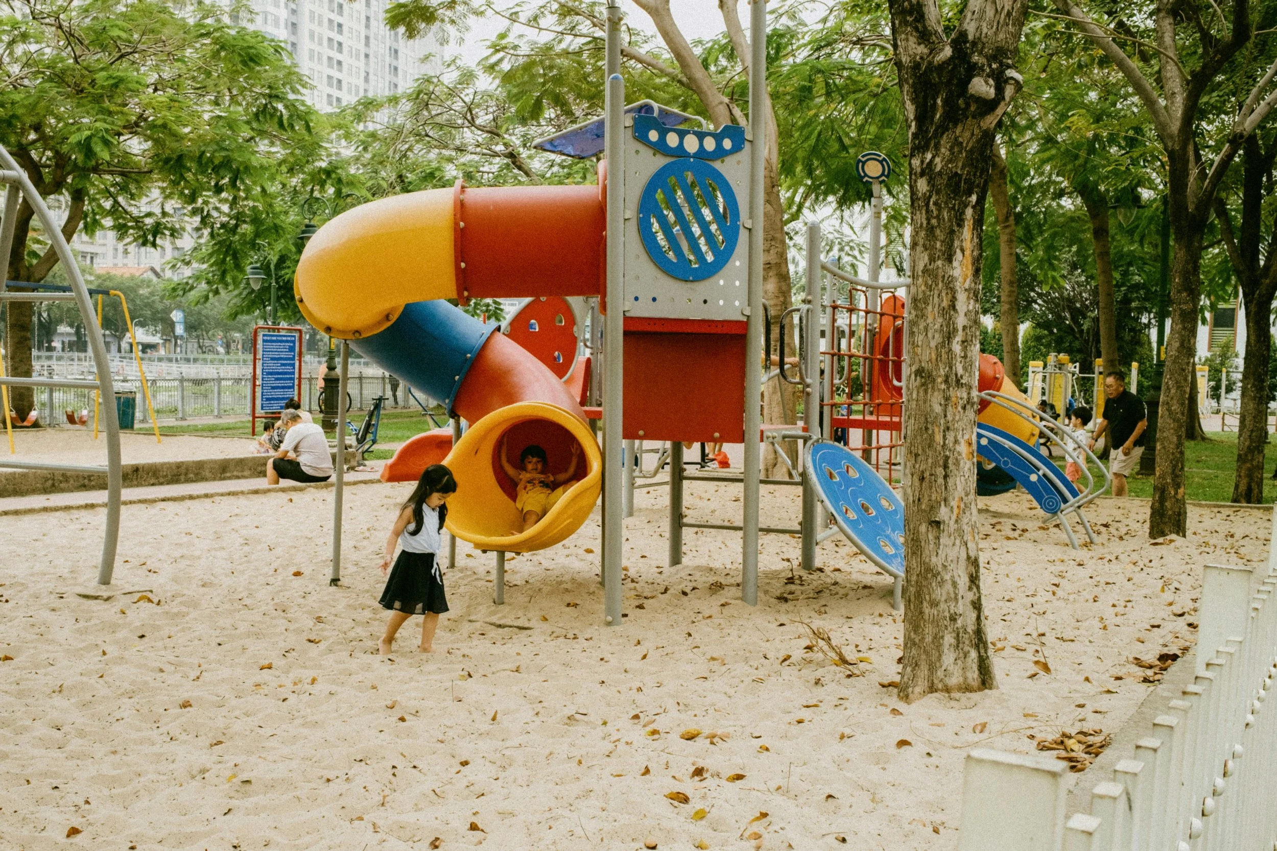 Children playing on a colorful playground slide and climbing structure in a park with trees, sand, and a cityscape in the background.