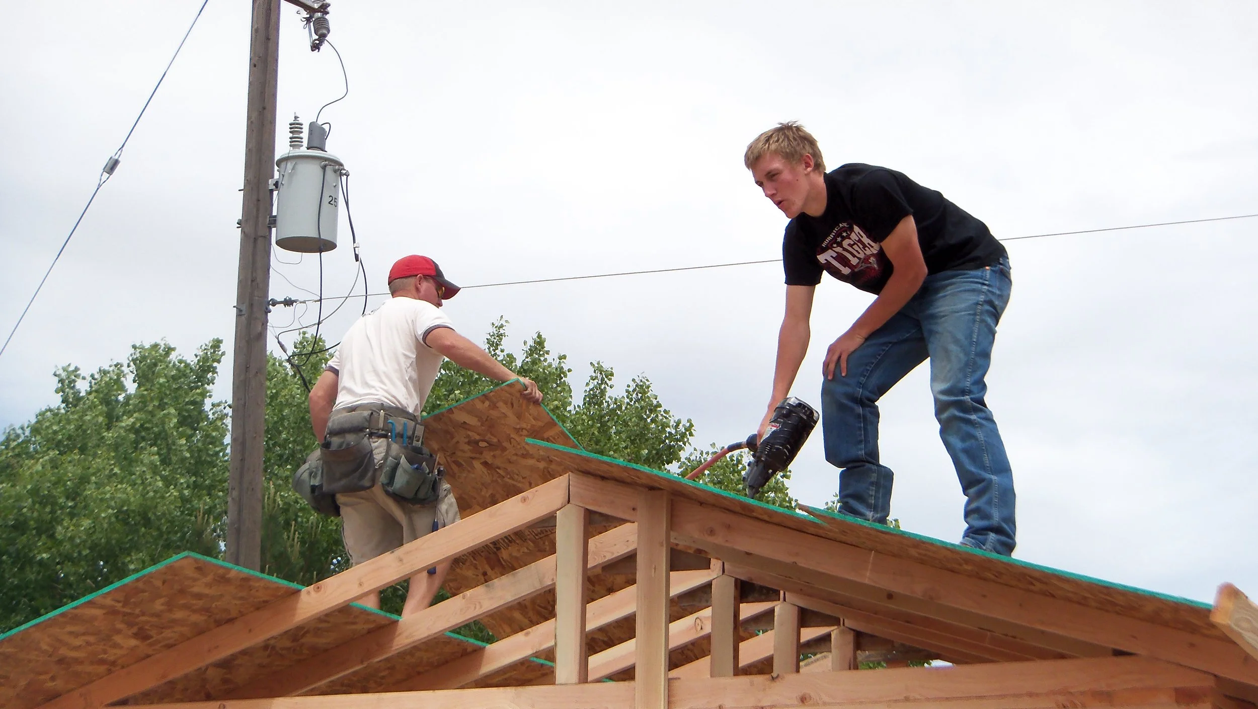 Two men working on a wooden structure, one wearing a white shirt and red cap, and the other in a black shirt, on top of the building. A large utility pole and trees are in the background.