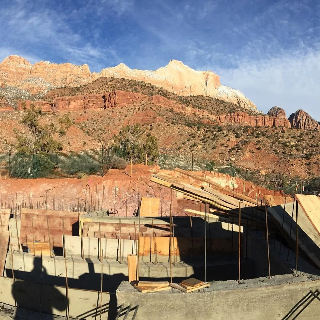 Construction site with concrete foundation and wooden planks against a backdrop of red and white rock mountains under a blue sky.