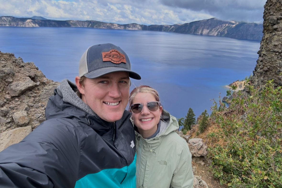 Smiling man and woman taking a selfie outdoors near Oregon's Crater Lake, with rugged rocks and green bushes around, and cloudy sky above.