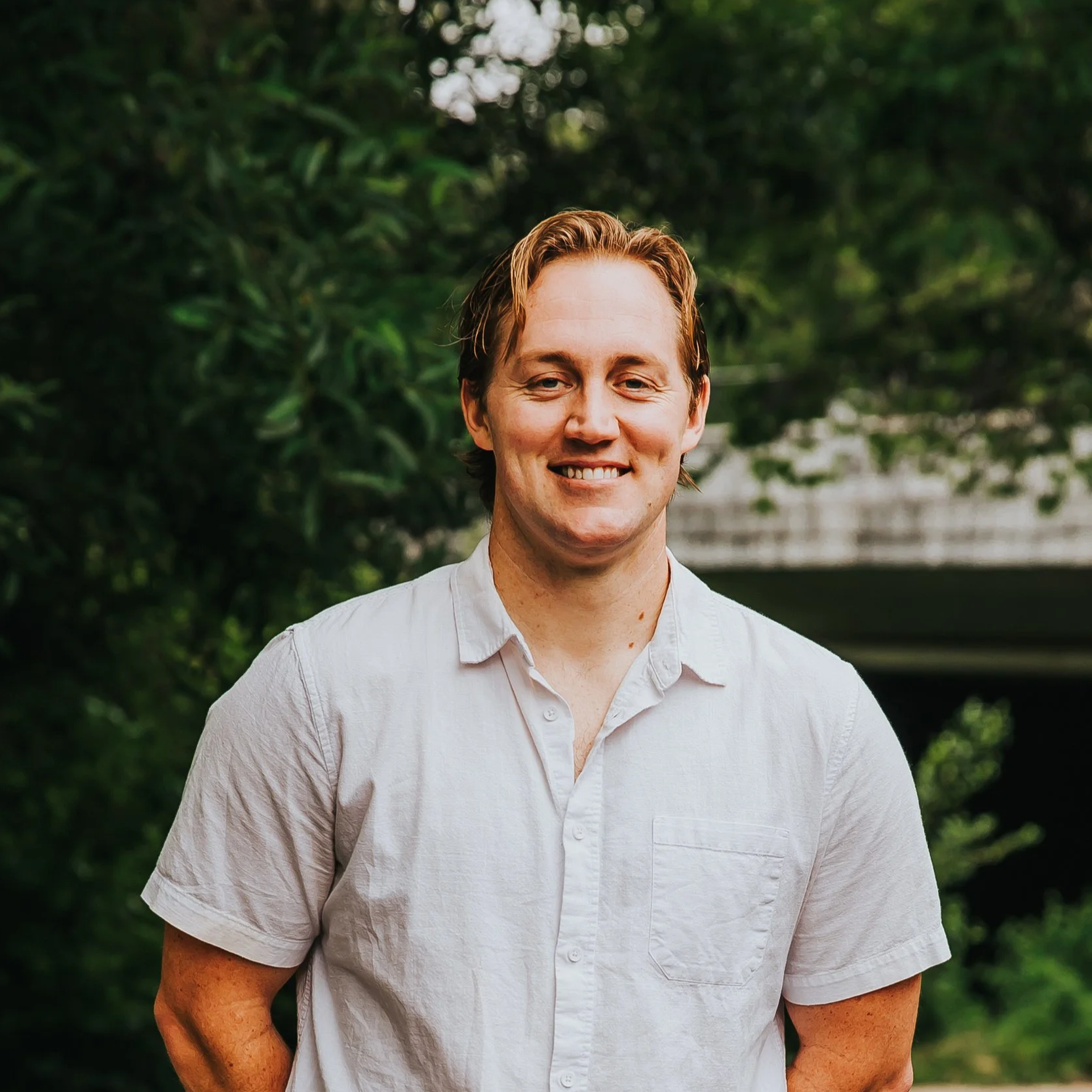 Portrait of the owner smiling with a white shirt and blonde medium hair green foliage in the background.