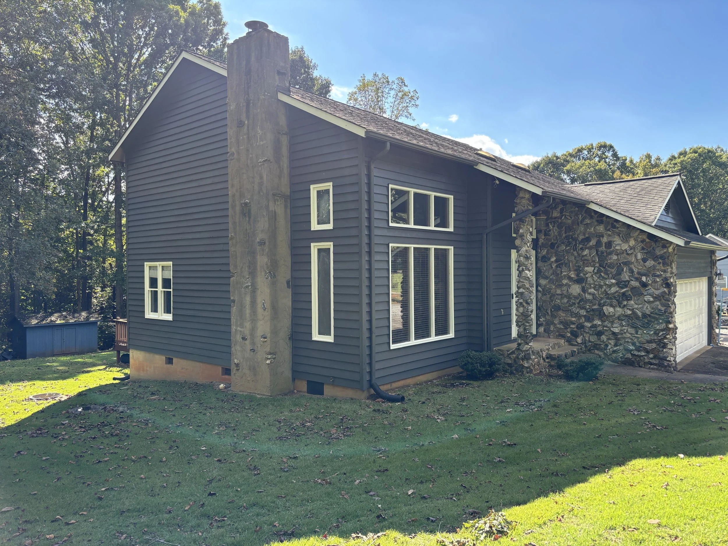 Side view of a house with dark gray siding, multiple windows, a stone chimney, and a garage, surrounded by a grassy yard and trees, on a sunny day.