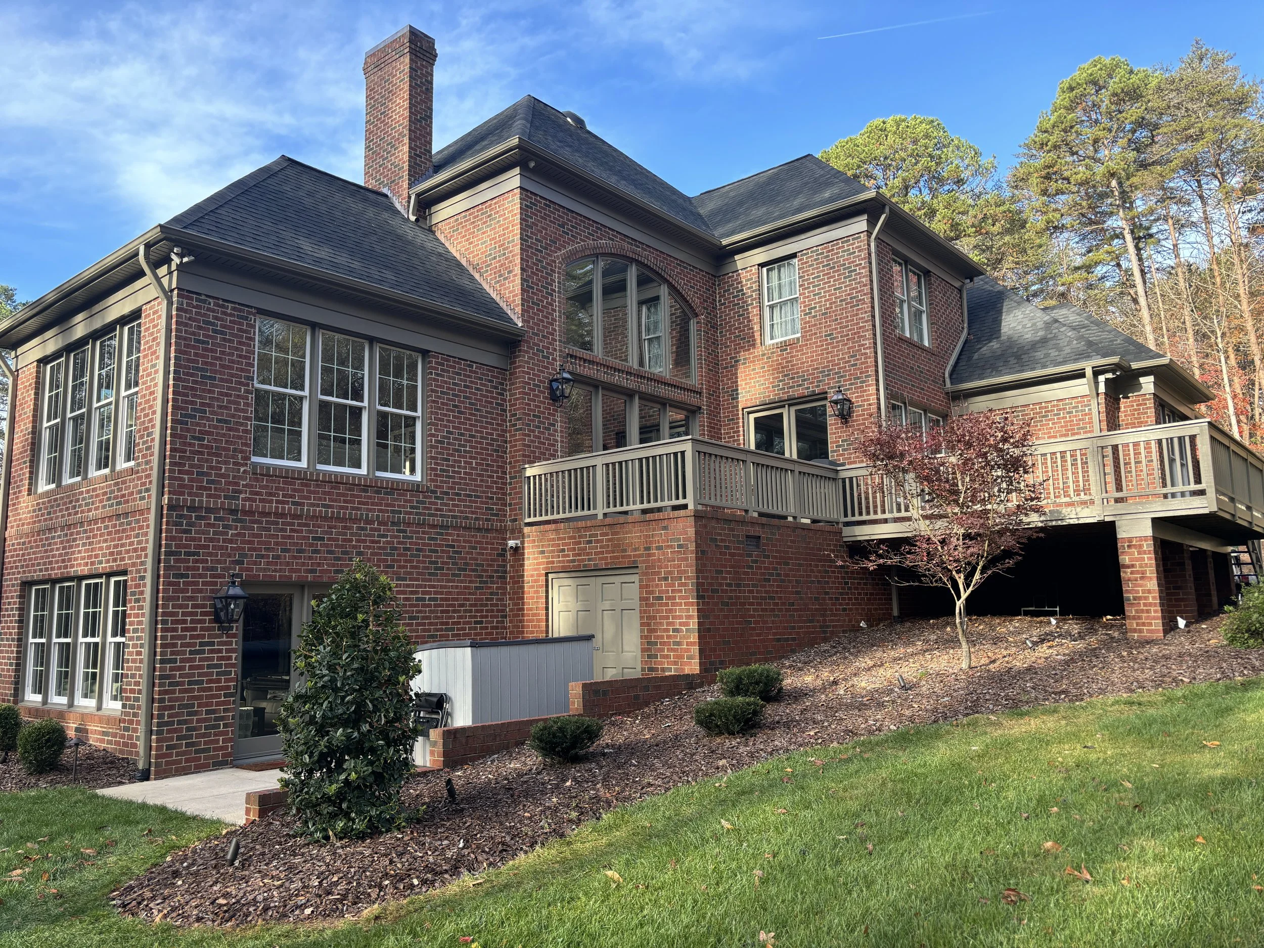 Large brick house with multiple windows, a balcony, and a sloped roof, surrounded by green lawn and trees, under a clear blue sky.