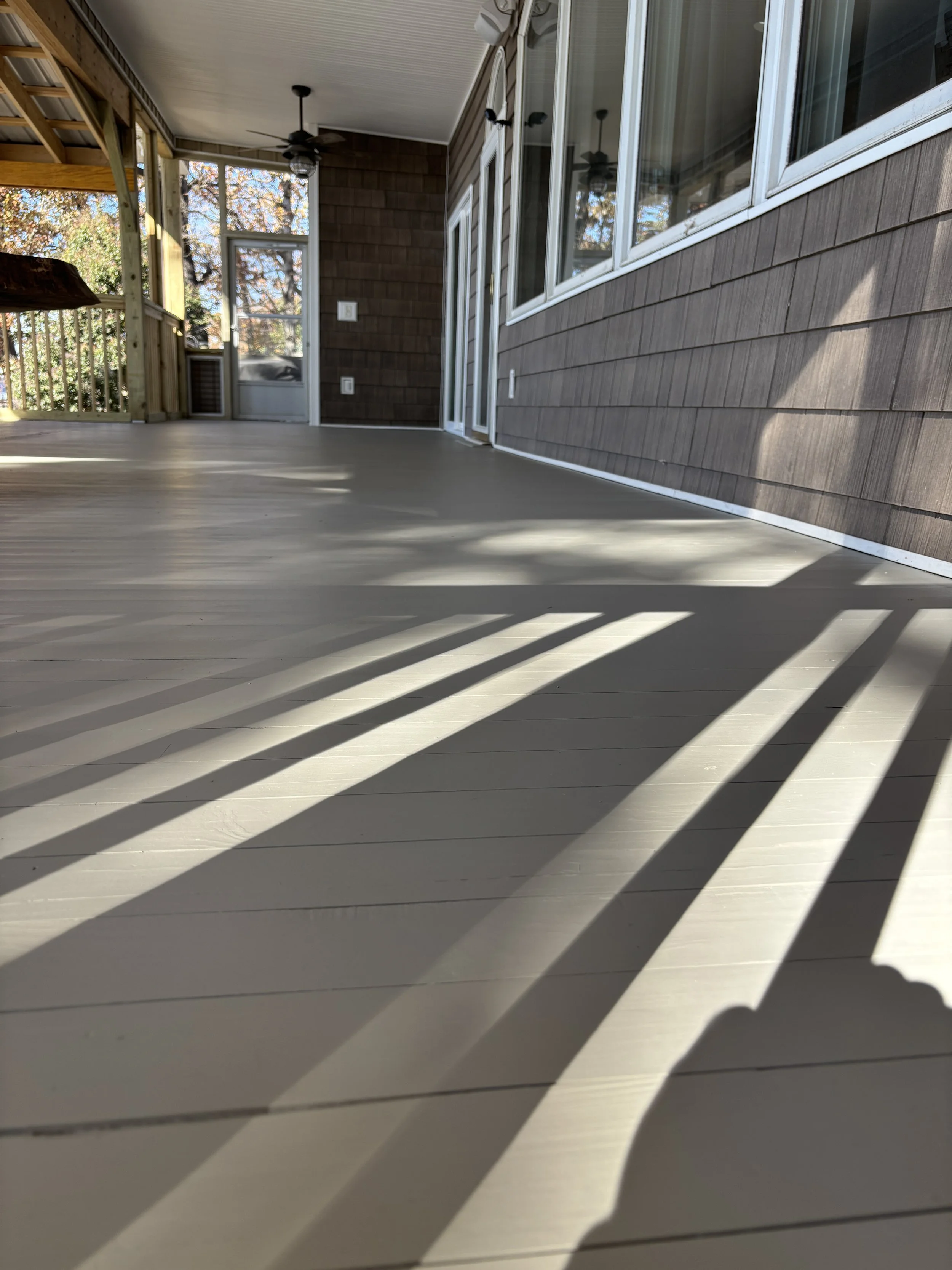 Photo of a covered porch with wooden flooring, shadow patterns from the railing, large sliding glass doors, and a ceiling fan.