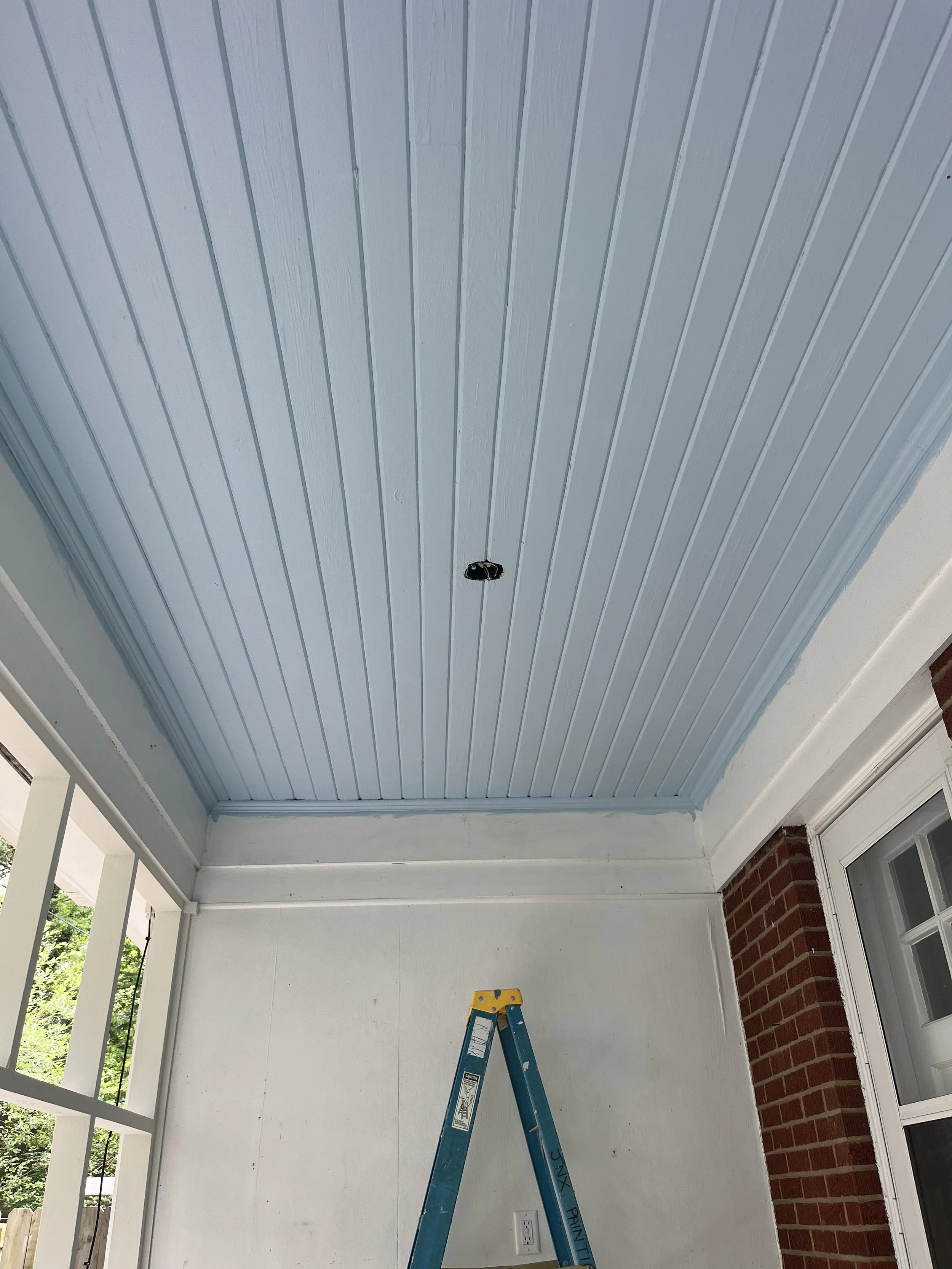 Ceiling with blue painted beadboard paneling, hole in ceiling, ladder inside a porch or sunroom with brick and window panes.