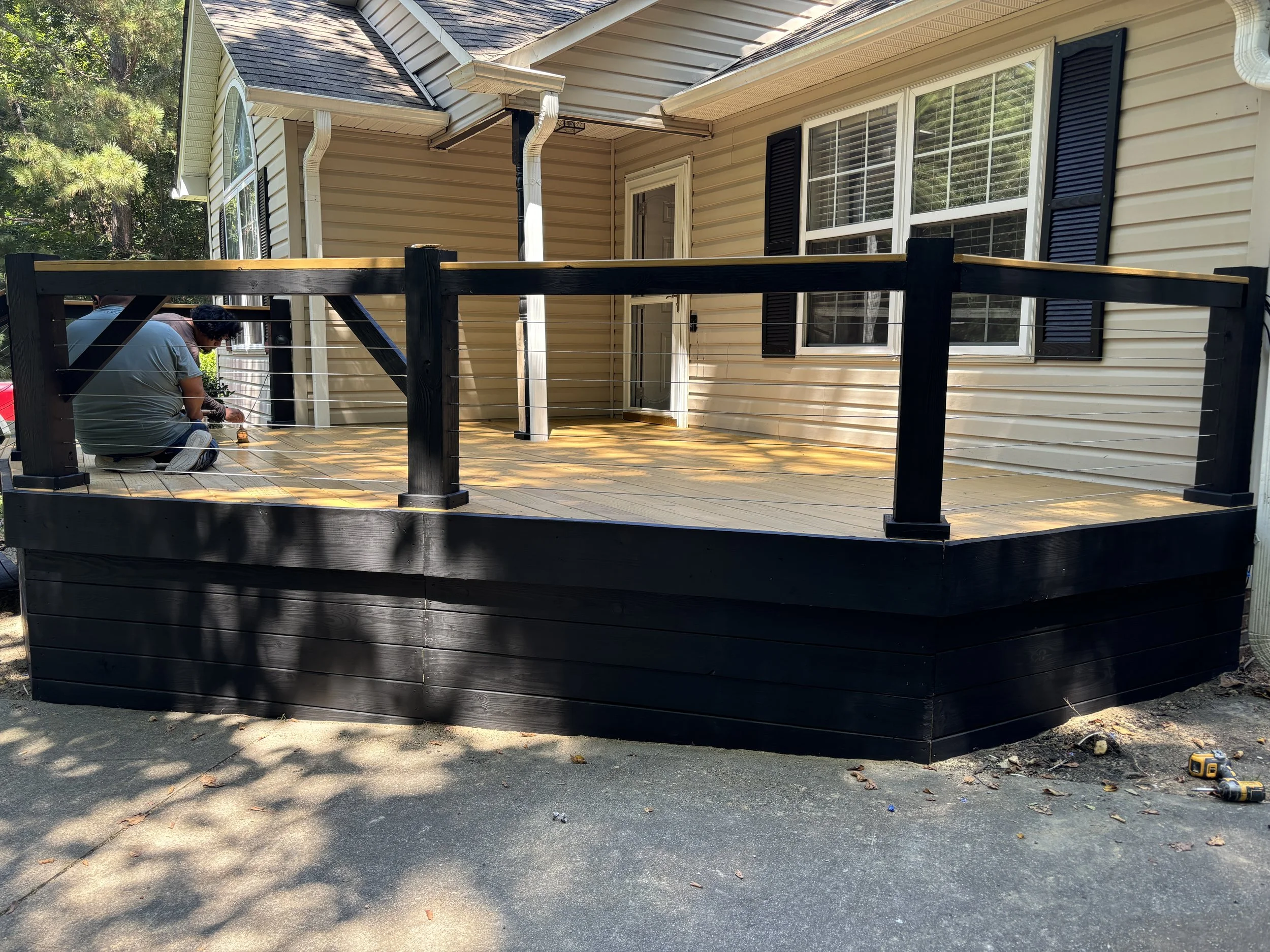 A person installing a wooden deck outside a house with beige siding and black shutters, surrounded by trees.