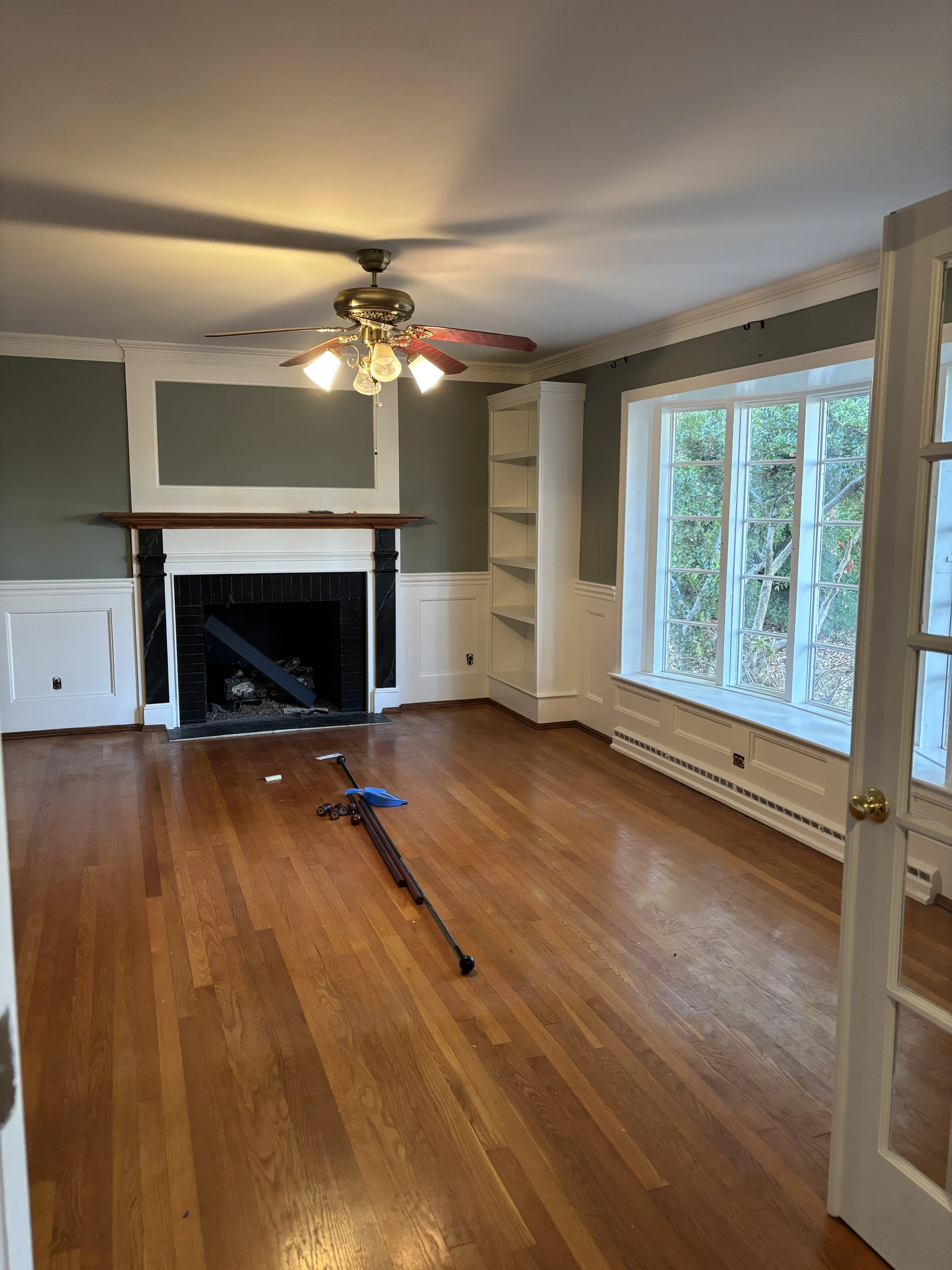 Empty living room with hardwood floors, a ceiling fan with lights, a fireplace with black tiles and a wood mantel, a large window, and built-in shelves.