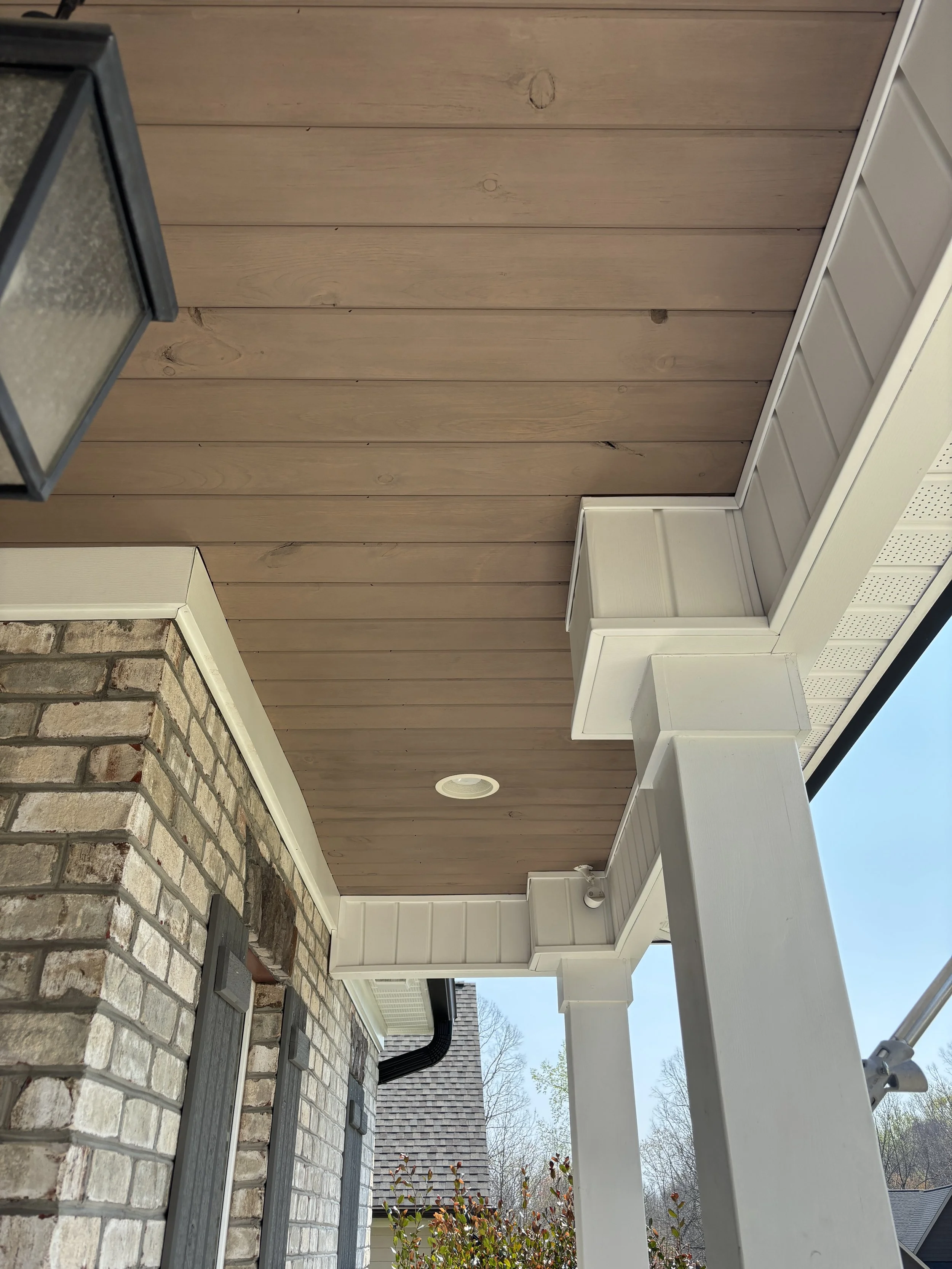 View of house porch ceiling with wooden paneling, white trim, brick wall, outdoor light fixture, and part of roof with sky and trees in background.