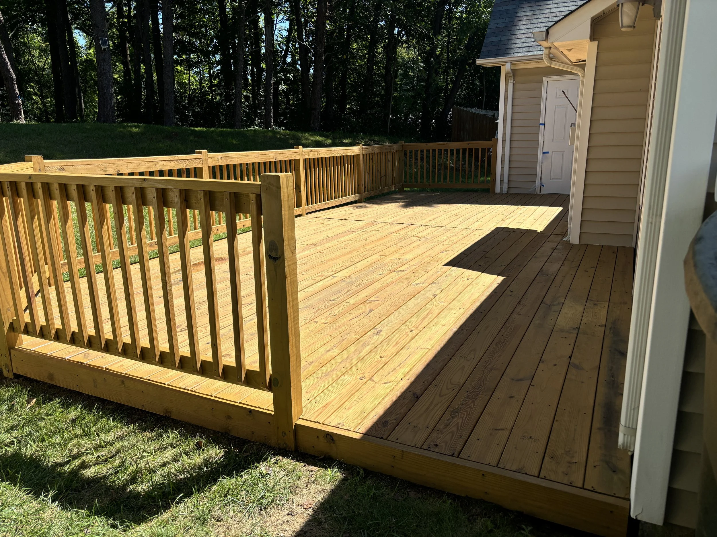 Newly built wooden deck attached to house with a small gate, surrounded by grass and a wooded area in the background.