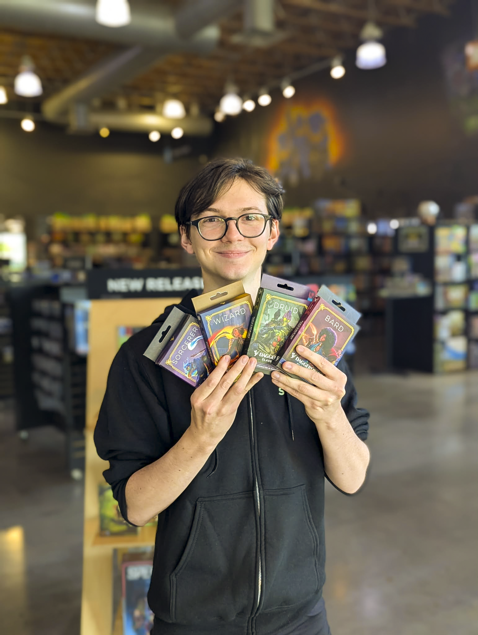 A person with short dark hair, glasses, and light skin smiling while holding multiple tabletop role-playing game packs in a library or game store.