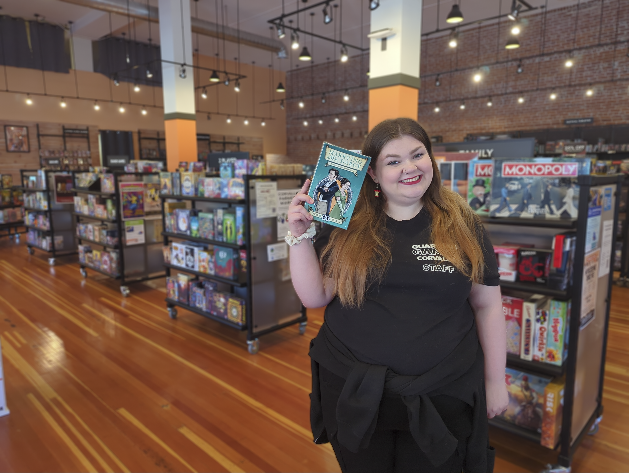 A young woman with long brown hair and a nose ring smiling as she holds a copy of the book 'Marrying Mr. Darcy' inside a bookstore. She is wearing a black shirt with text and necklace earrings shaped like flags. The bookstore has shelves filled with books and a brick wall background.