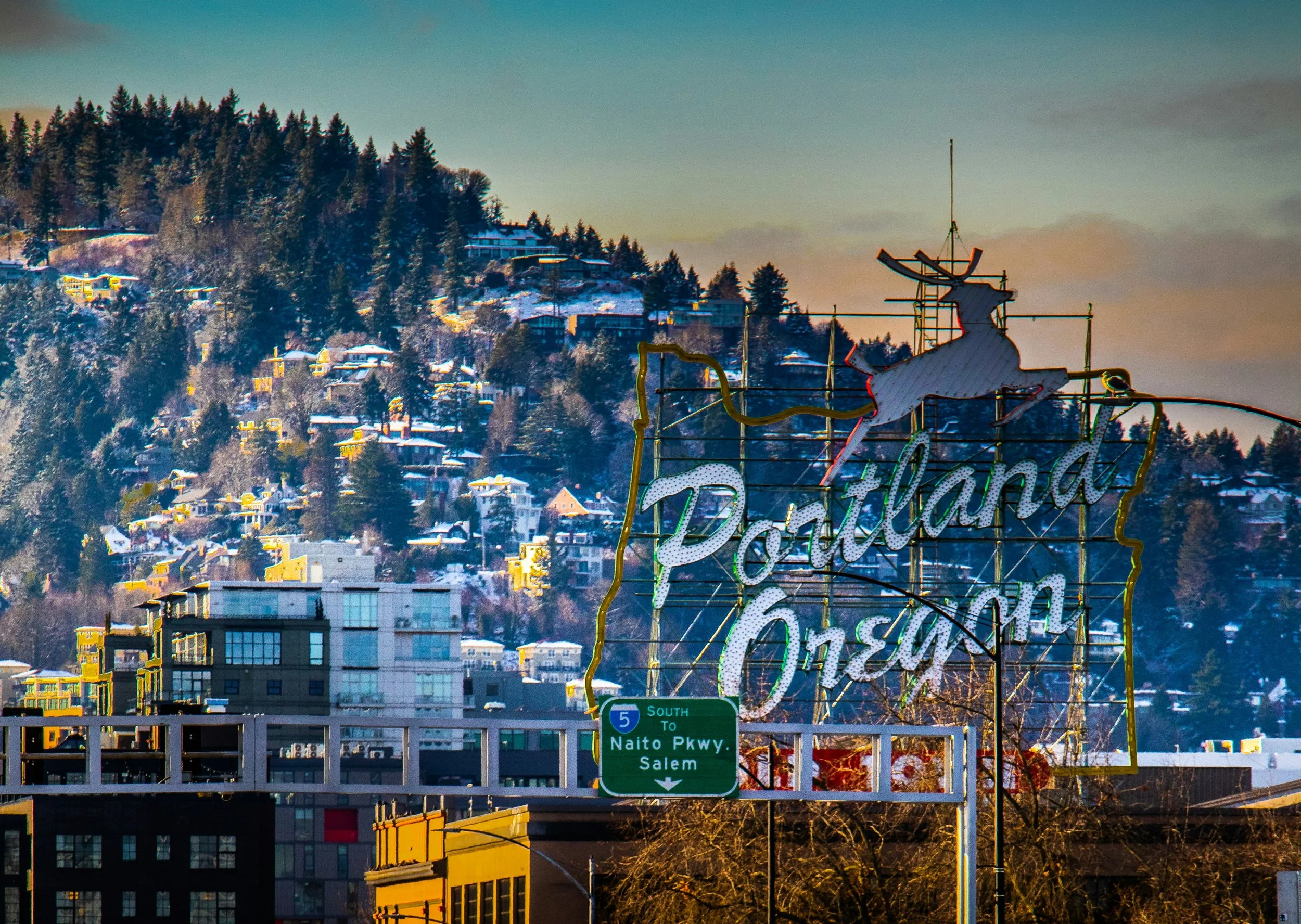 A cityscape with residential buildings on a snowy hillside, a neon Bing Crosby sign, and a highway sign indicating southbound directions to Naito Parkway and Salem.