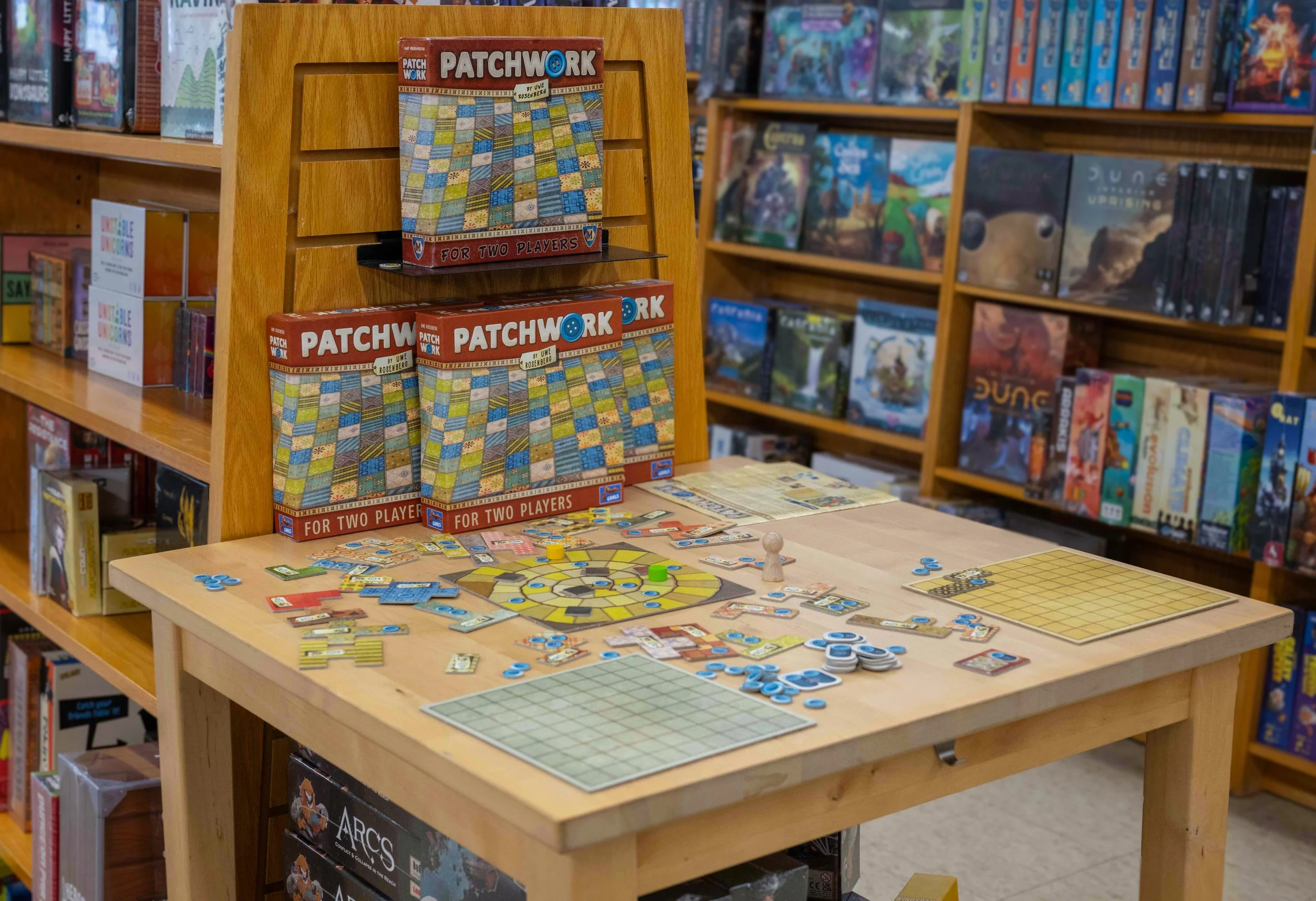 A display of the board game Patchwork on a wooden shelf in a bookstore or game shop, with other books and games on shelves in the background.