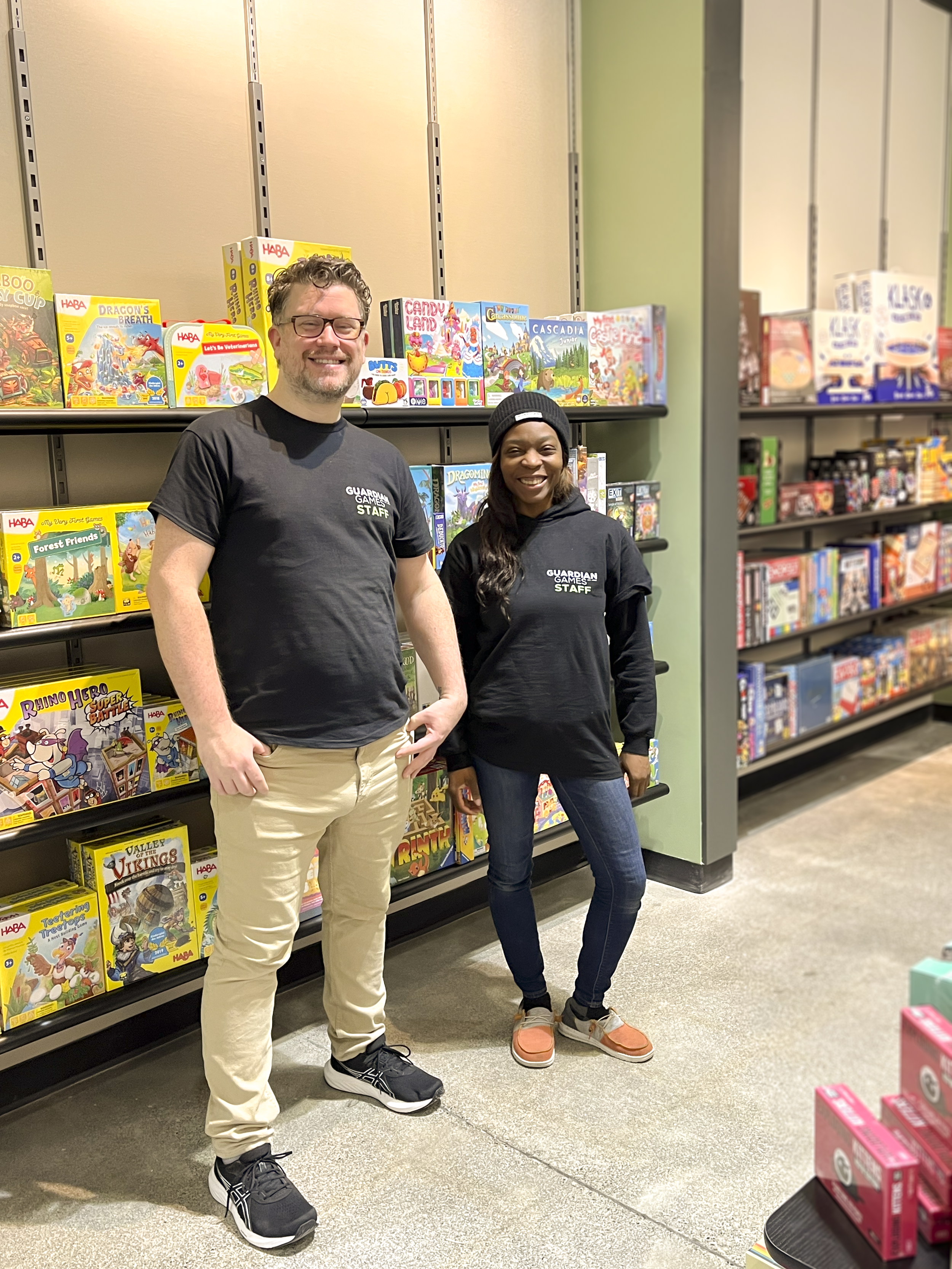 Two people standing in front of a shelf of board games in a retail store, smiling at the camera. The person on the left is a man with glasses, wearing a black Guardian Games staff T-shirt and beige pants. The person on the right is a woman with long dark hair, wearing a gray beanie, a black Guardian Games staff T-shirt, and jeans.