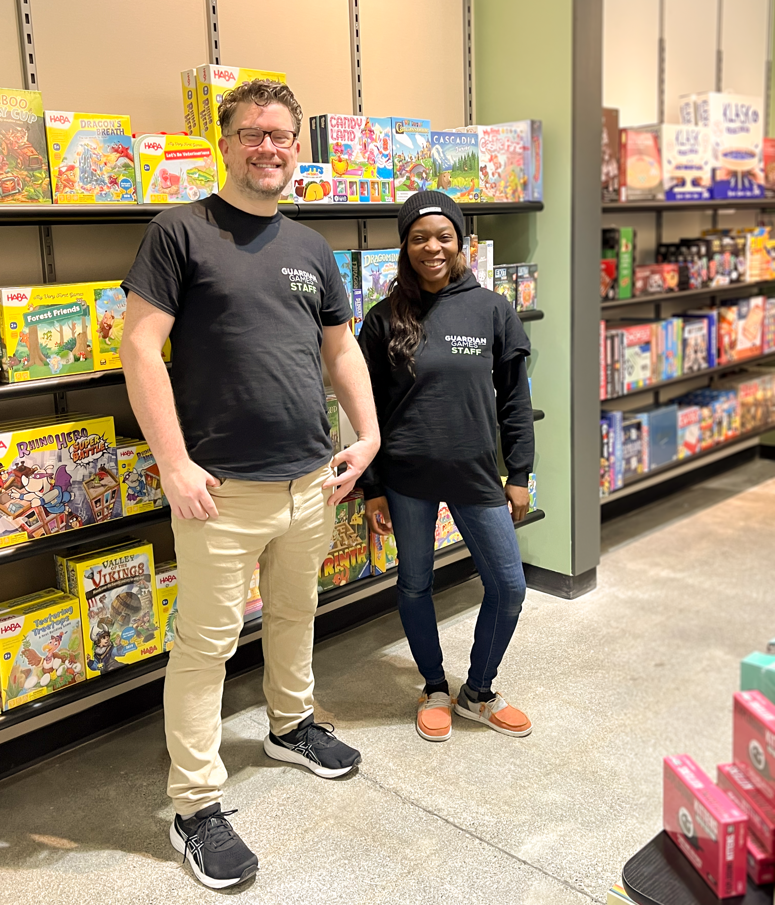 Two people, one man and one woman, standing inside a game and toy store, smiling at the camera. The man is wearing glasses, a black Guardian Games staff T-shirt, khaki pants, and black sneakers. The woman is wearing a black Guardian Games staff hoodie, dark jeans, and orange sneakers. Behind them are shelves filled with children's board games and puzzles.
