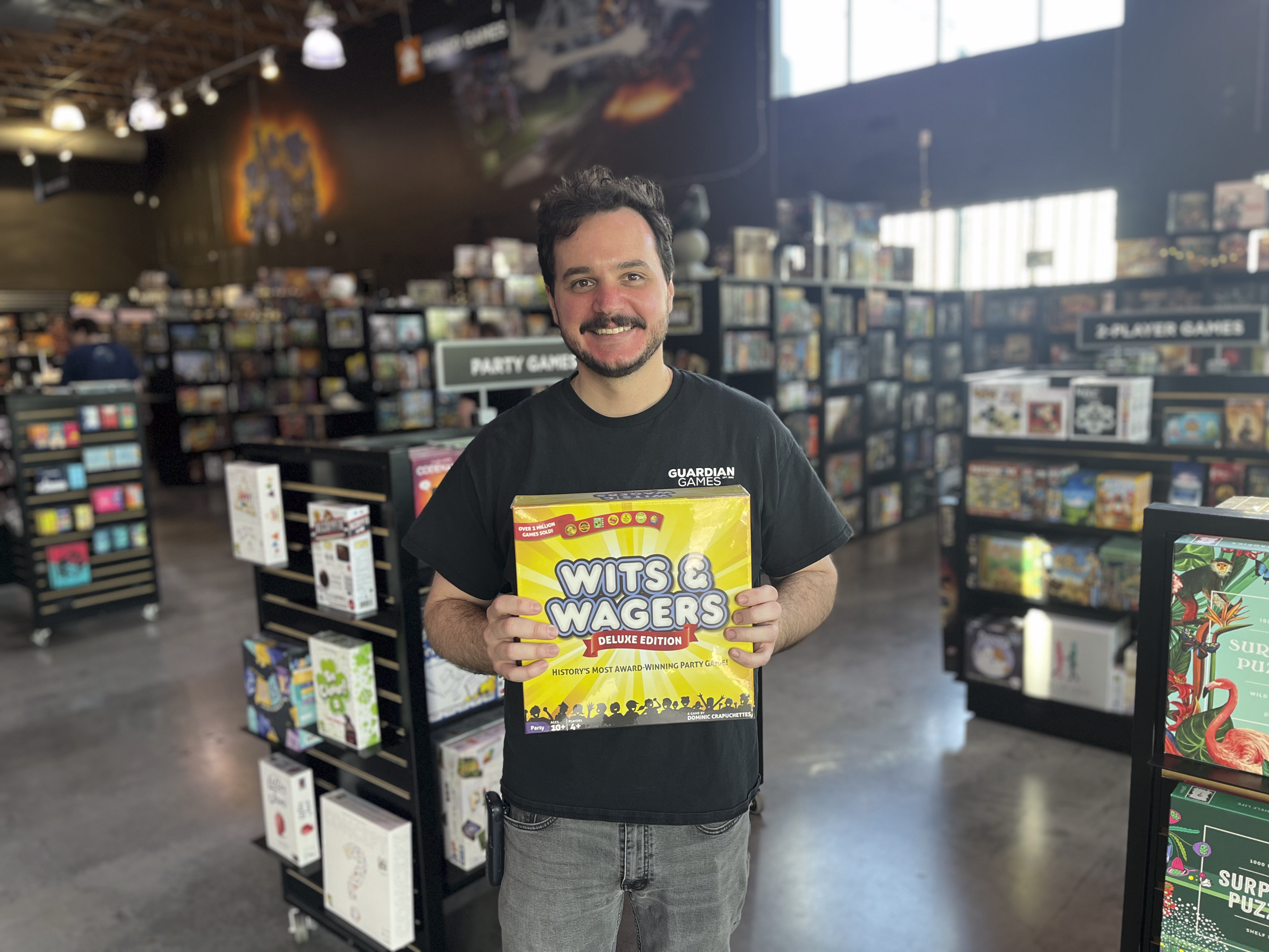 A man with dark hair and a beard smiling at the camera inside a game store while holding a box of Wits & Wagers deluxe edition board game. The store has shelves filled with various board games and is well-lit with natural and artificial lighting.