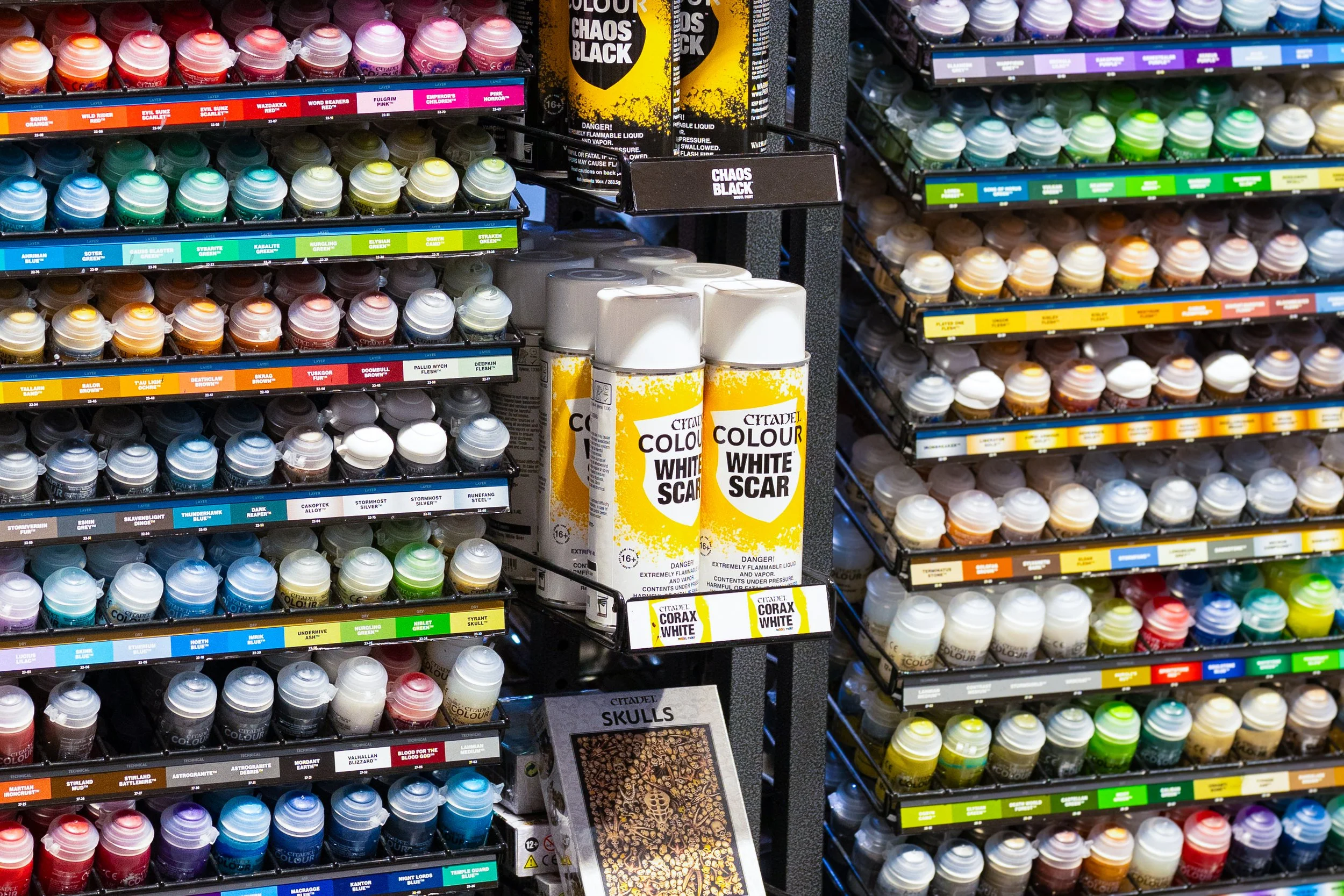 Shelf display of various bottles of Citadel paint for miniature modeling, with labels indicating different colors and finishes, including chaos black and white scar, alongside some other hobby-related supplies.