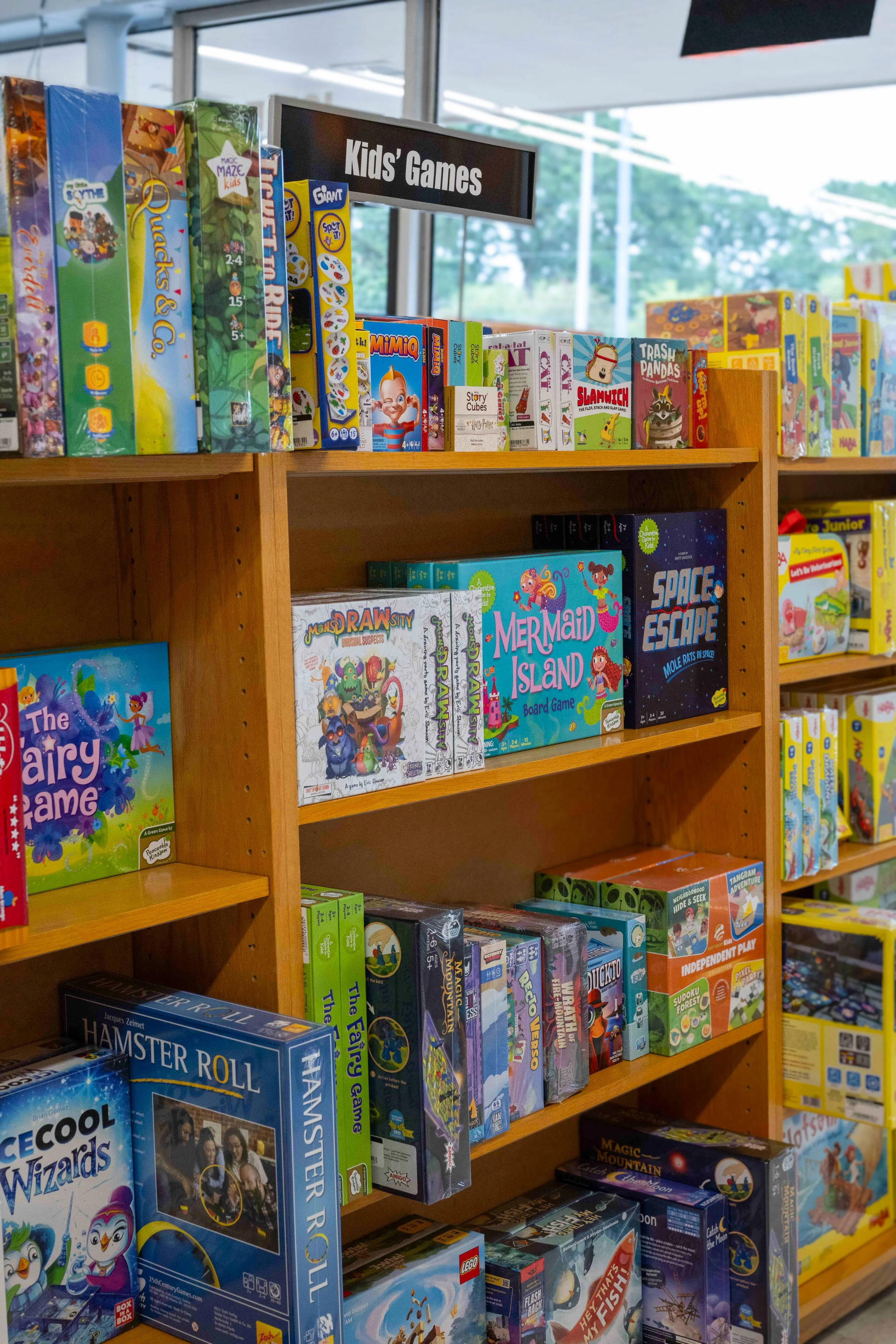 A wooden shelf filled with various children's board games and puzzles in a store, labeled 'Kids' Games' on a sign above.