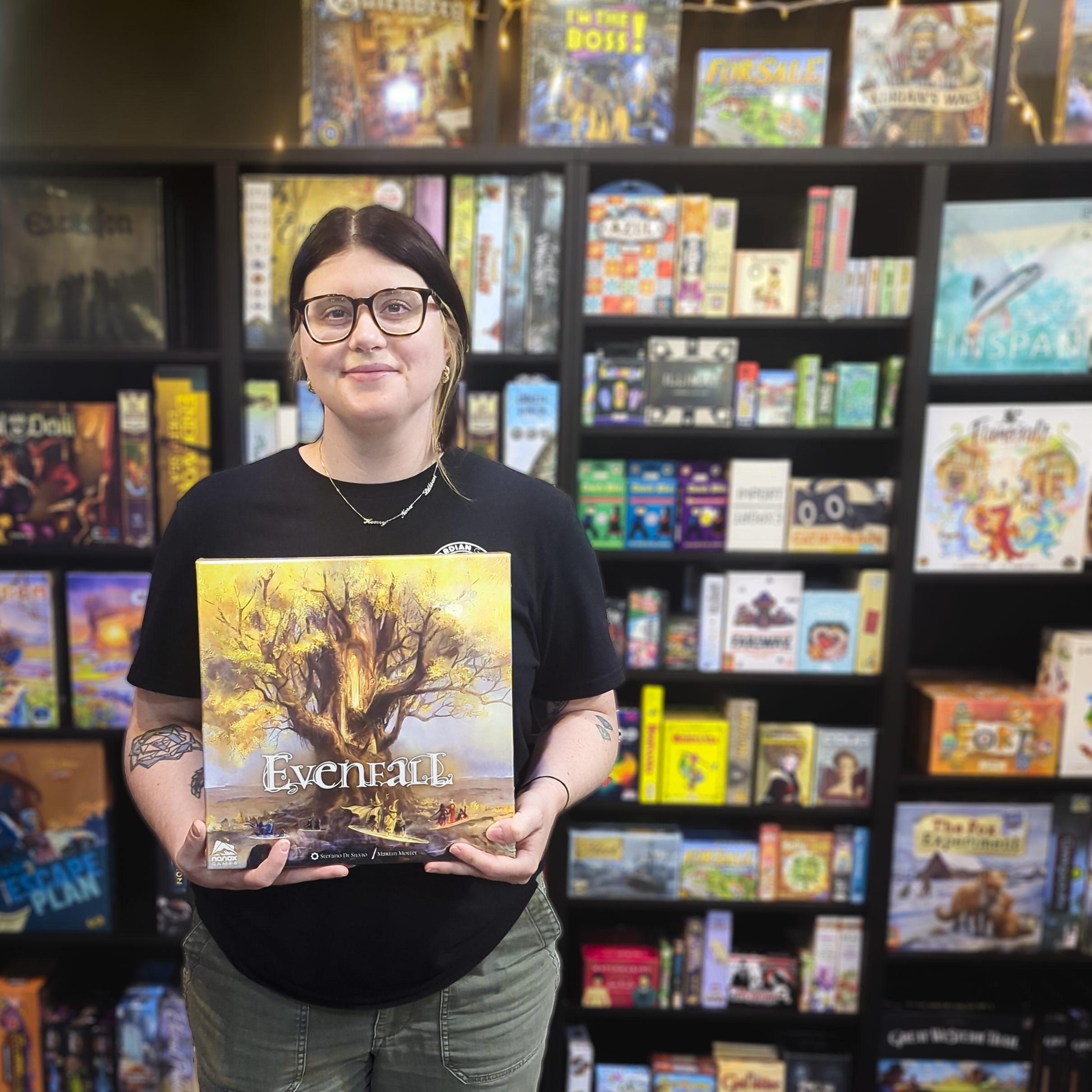 A woman with black hair, glasses, and tattoos on her arm is holding a board game box titled 'Everdell' in a bookstore. She is wearing a black shirt with a logo and gray pants. Behind her are shelves filled with colorful board games.