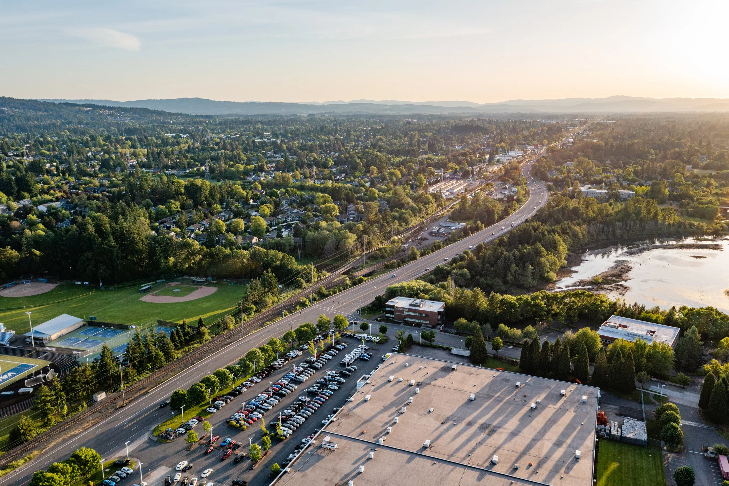 An aerial view of a suburban area at sunset, showing a highway with cars, a parking lot filled with vehicles, a baseball field, tennis courts, trees, residential houses, and a body of water.