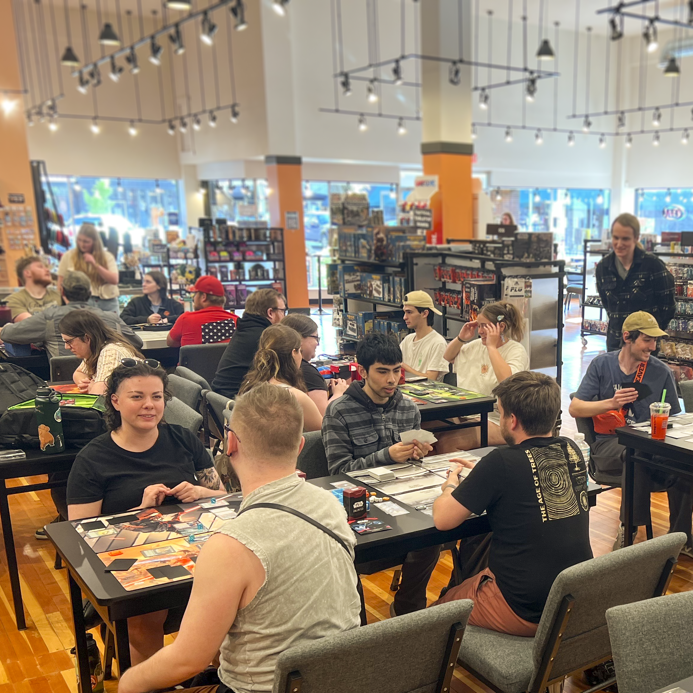 People playing and spectating a tabletop game event inside a store, with shelves of games and large windows in the background.