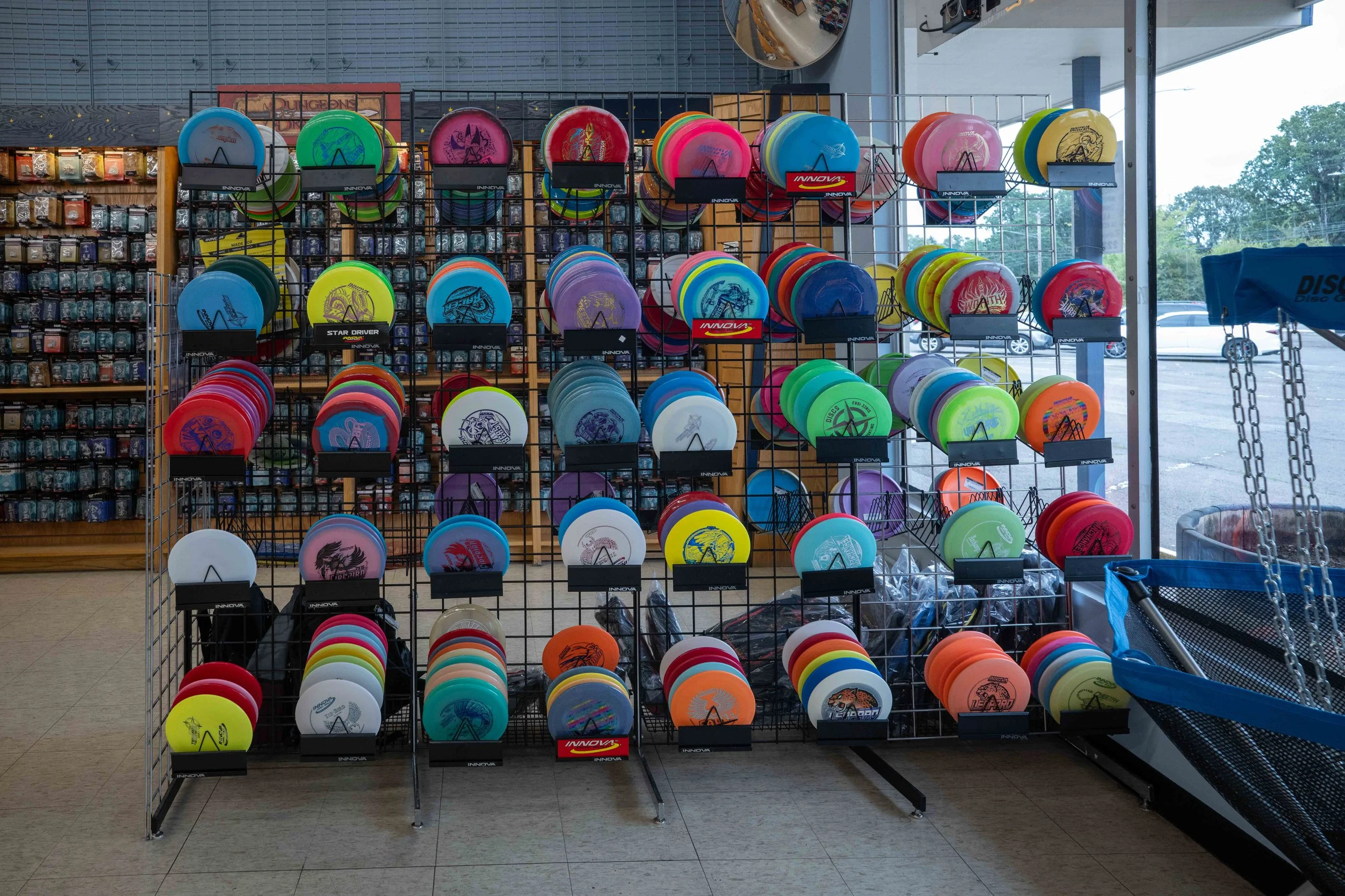 Display of colorful frisbees on a wire rack inside a store, with a window revealing a parking lot and trees outside.