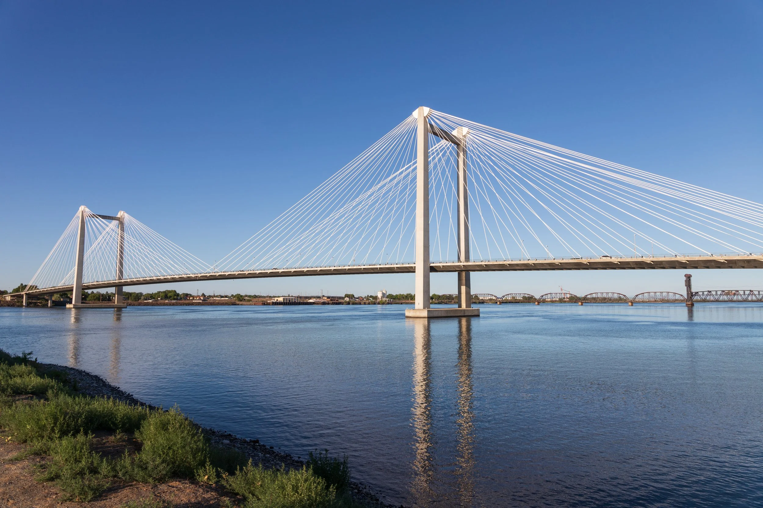 A modern cable-stayed bridge over a body of water with clear blue sky in the background. The bridge has tall pylons and white cables, and there is a shoreline with green vegetation in the foreground.
