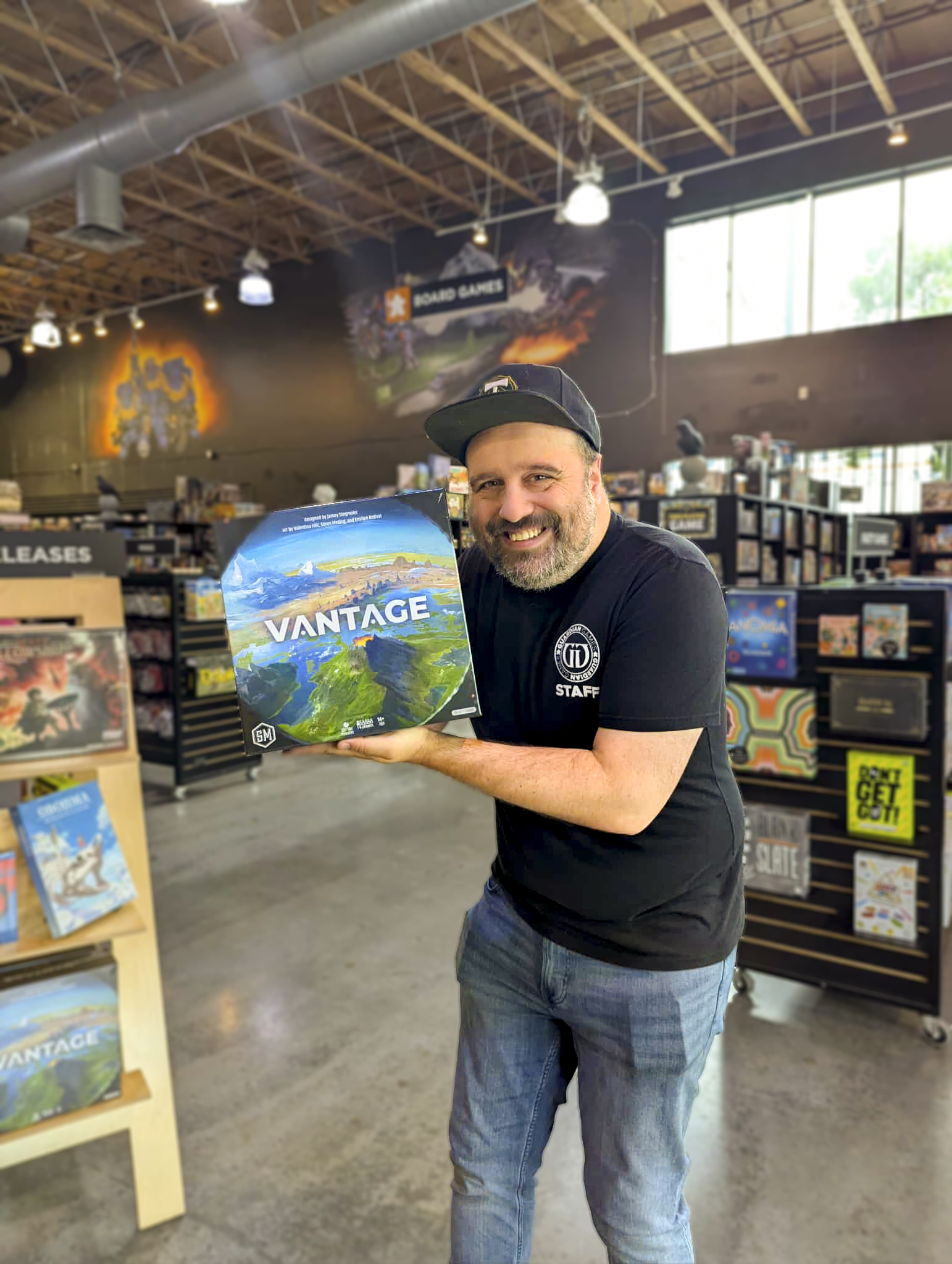 A man with a beard and black cap smiling and holding a board game titled 'VANTAGE' inside a game store with shelves of board games around him.