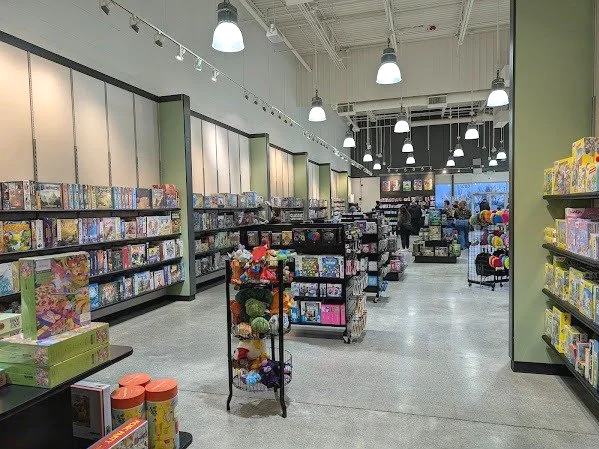 Inside a store with shelves of board games and toys, lit by hanging ceiling lights, with a few people browsing.