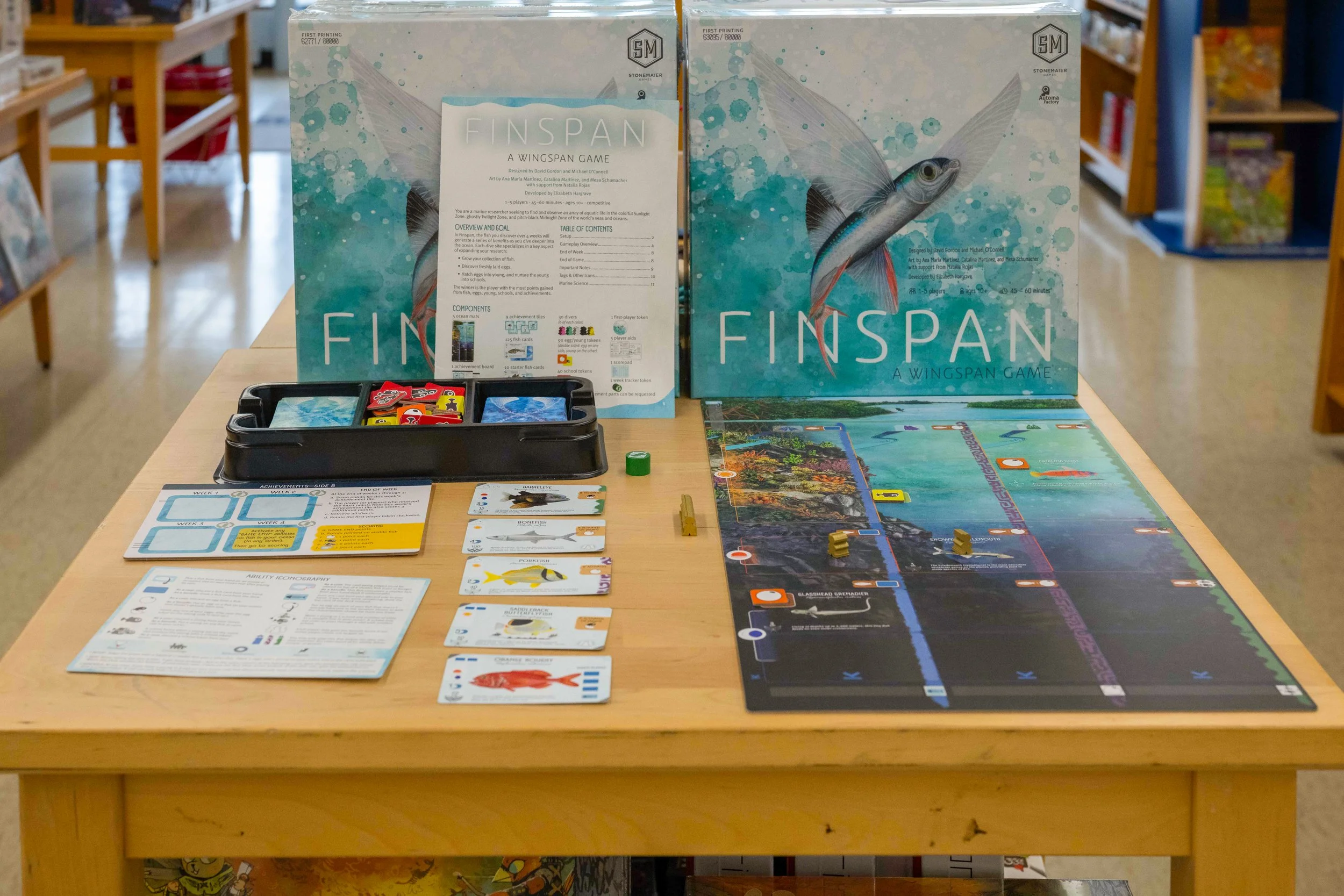 Board game titled Finspn on a store display table with game pieces, cards, and instructions, with shelves of other games in the background.