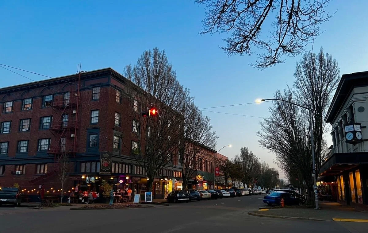 City street scene during dusk with a red traffic light, parked cars, and buildings with storefronts and outdoor dining.