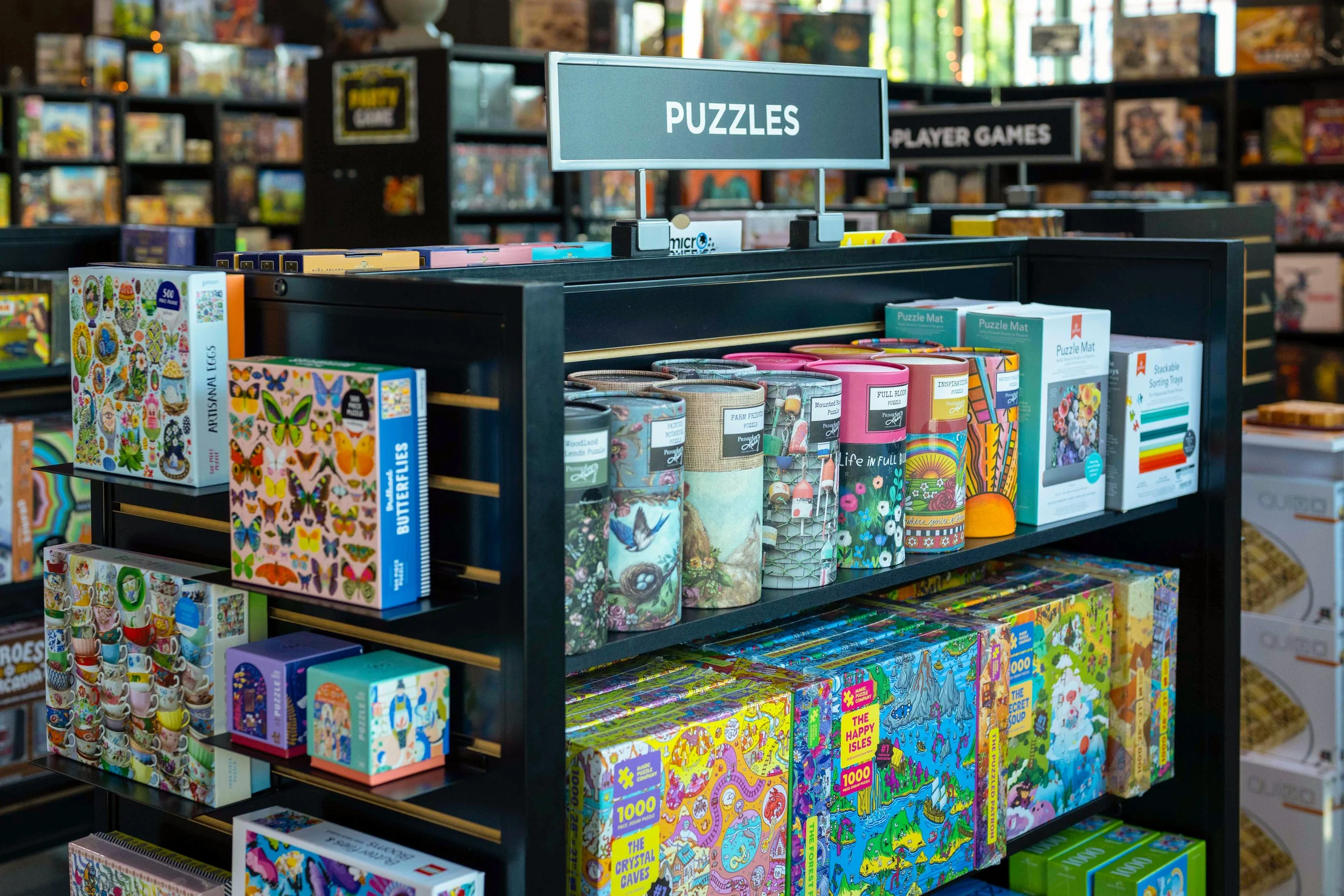 Store shelves displaying puzzle books, with signs labeled 'PUZZLES' and 'PLAYER GAMES' in a bookstore or game shop.