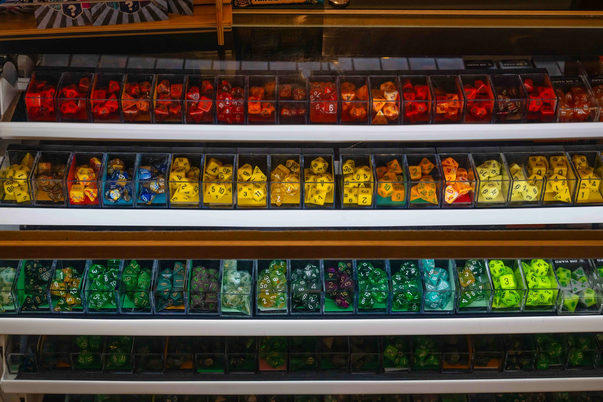 Display of various sets of multi-sided dice in clear plastic containers, organized by color: red, yellow, green, and purple, on store shelves.