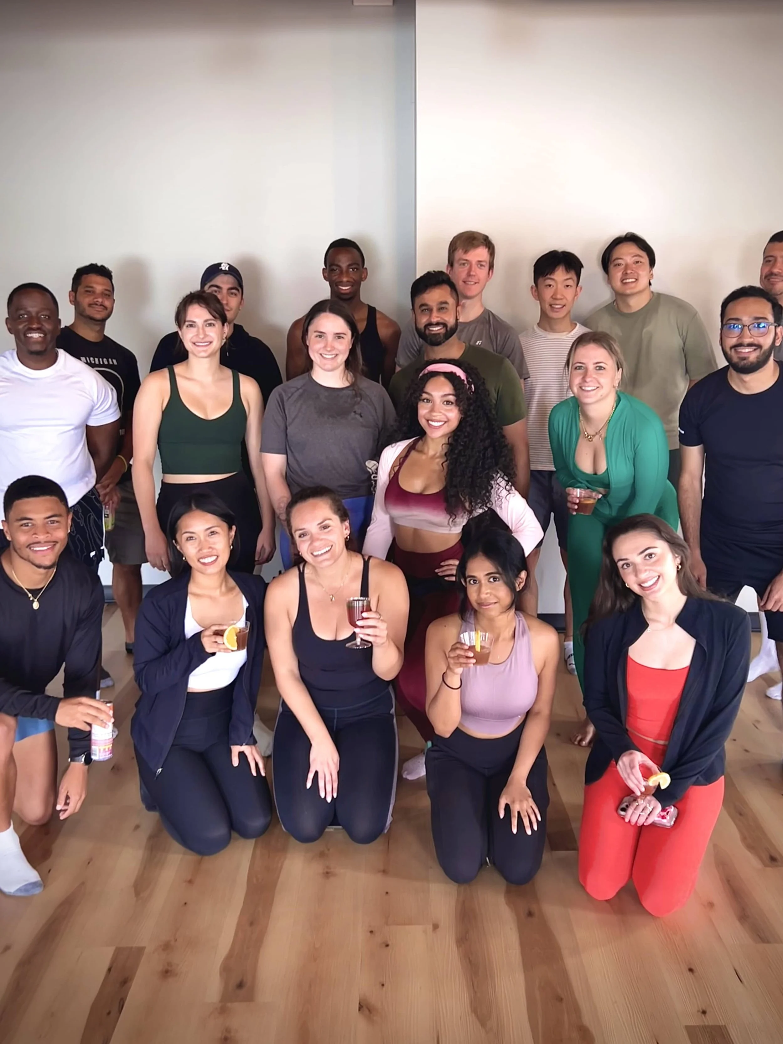 Group of diverse smiling people in casual attire, some holding drinks, posing indoors on a wooden floor with a plain wall background.