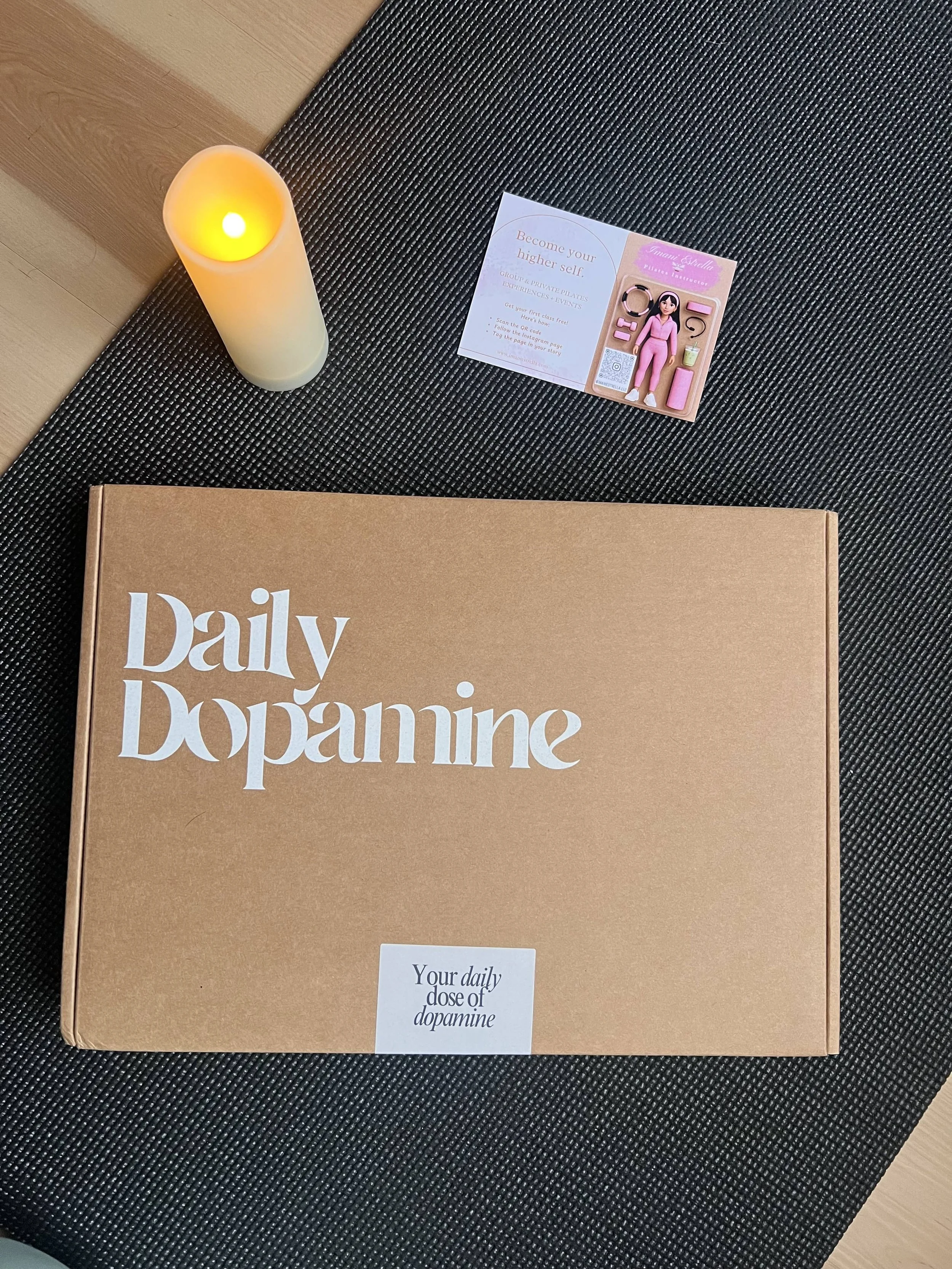 A brown box labeled "Daily dopamine" on a black textured surface with a candle, a business card, and a pink action figure with accessories next to it.