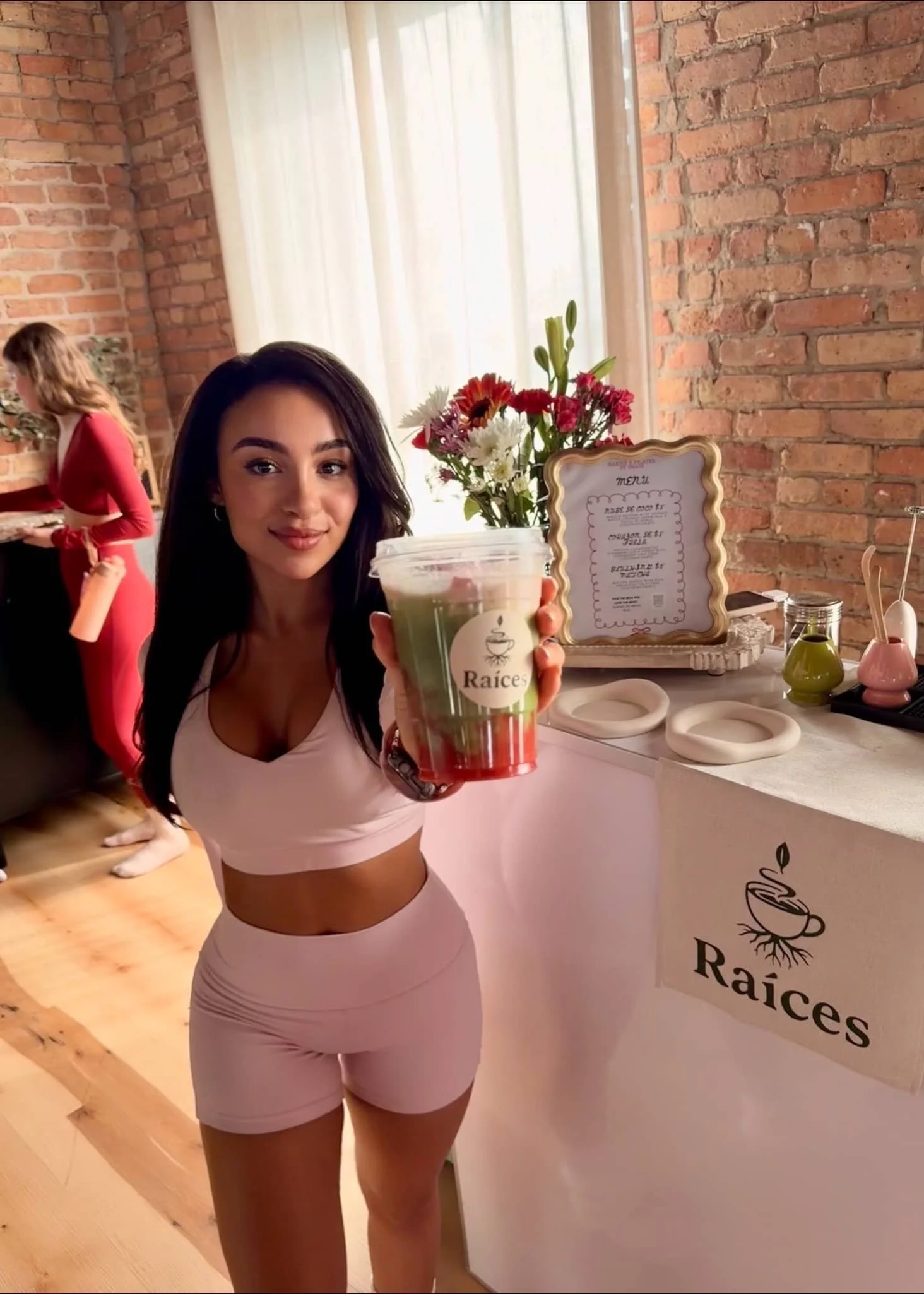 A woman in workout clothes holding a smoothie with a Raices logo, standing in a cafe with a brick wall, flowers, and a menu display in the background.