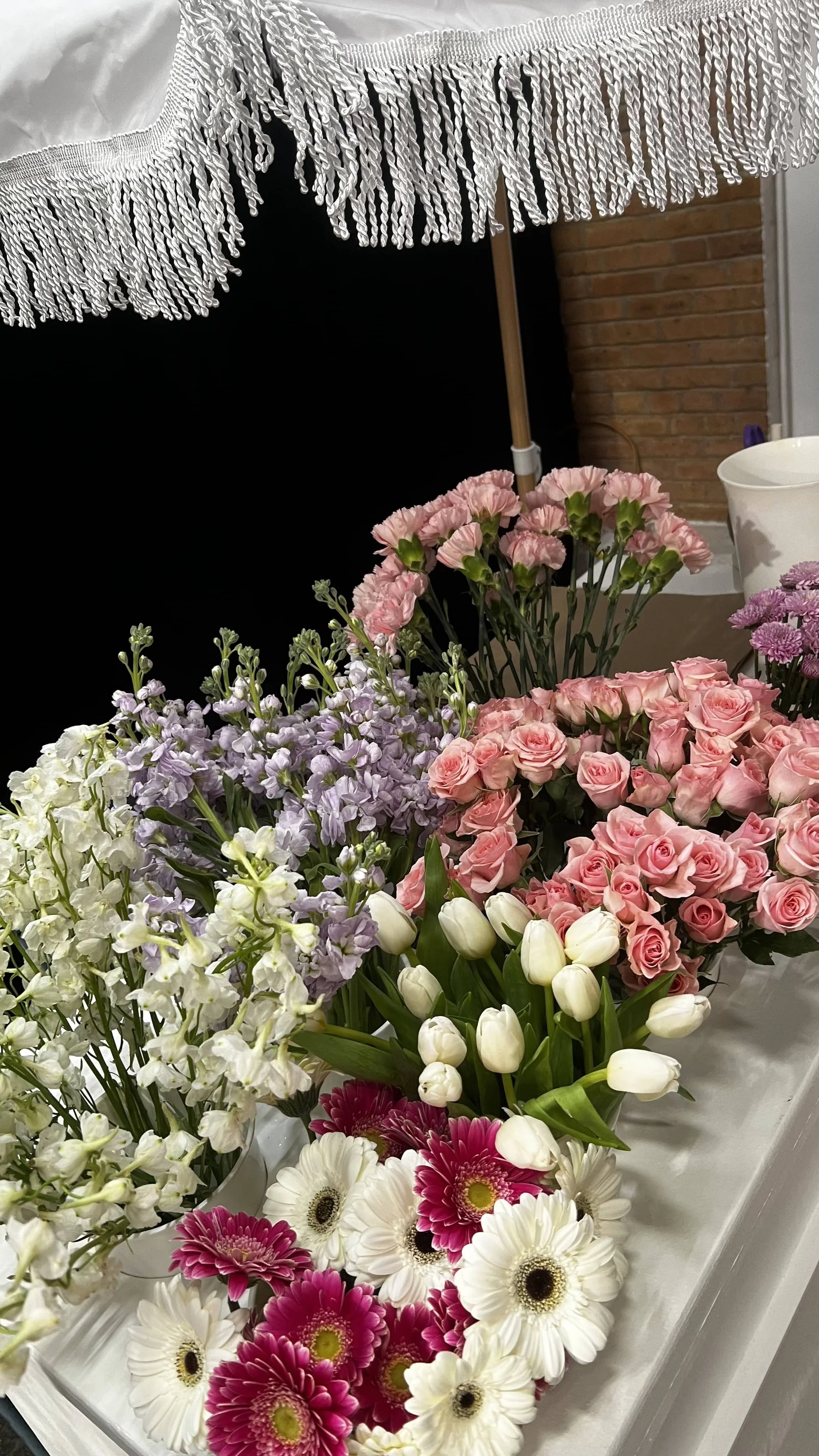 A variety of fresh flowers including pink roses, white tulips, white and pink daisies, purple stock, and light purple stock, arranged in vases on a white surface, with a brick wall and a patio umbrella in the background.