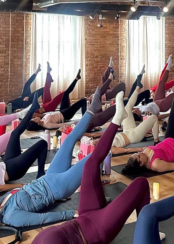 Group of women participating in a yoga class, lying on mats, raising one leg while supporting their heads with hands, in a studio with brick walls and large windows with curtains.
