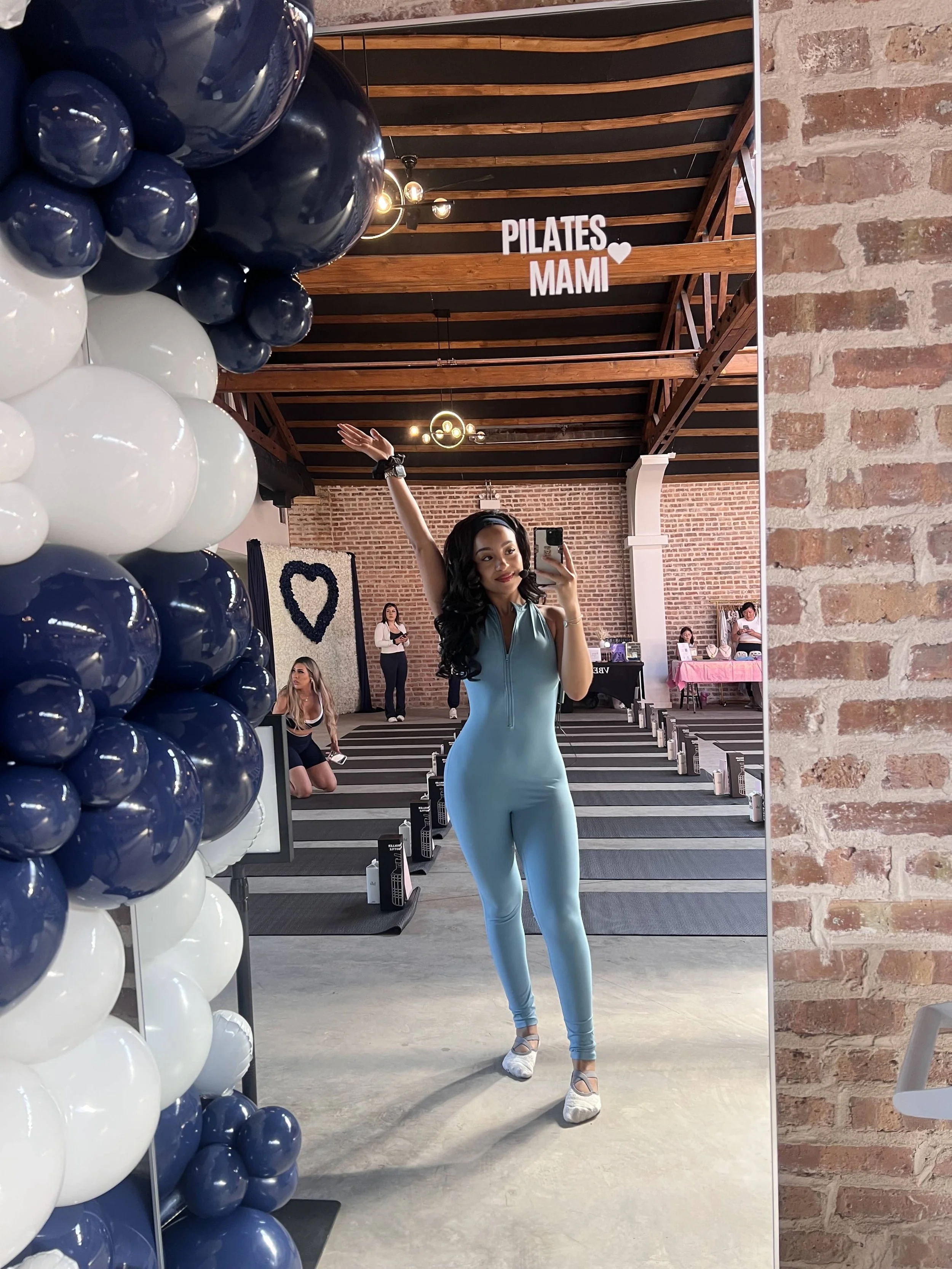 Young woman in a blue workout jumpsuit taking a mirror selfie at a fitness class. The background includes balloons in white and navy blue colors, a brick wall, and a sign that reads "Pilates Mami" with a heart. Other people are seated and preparing i